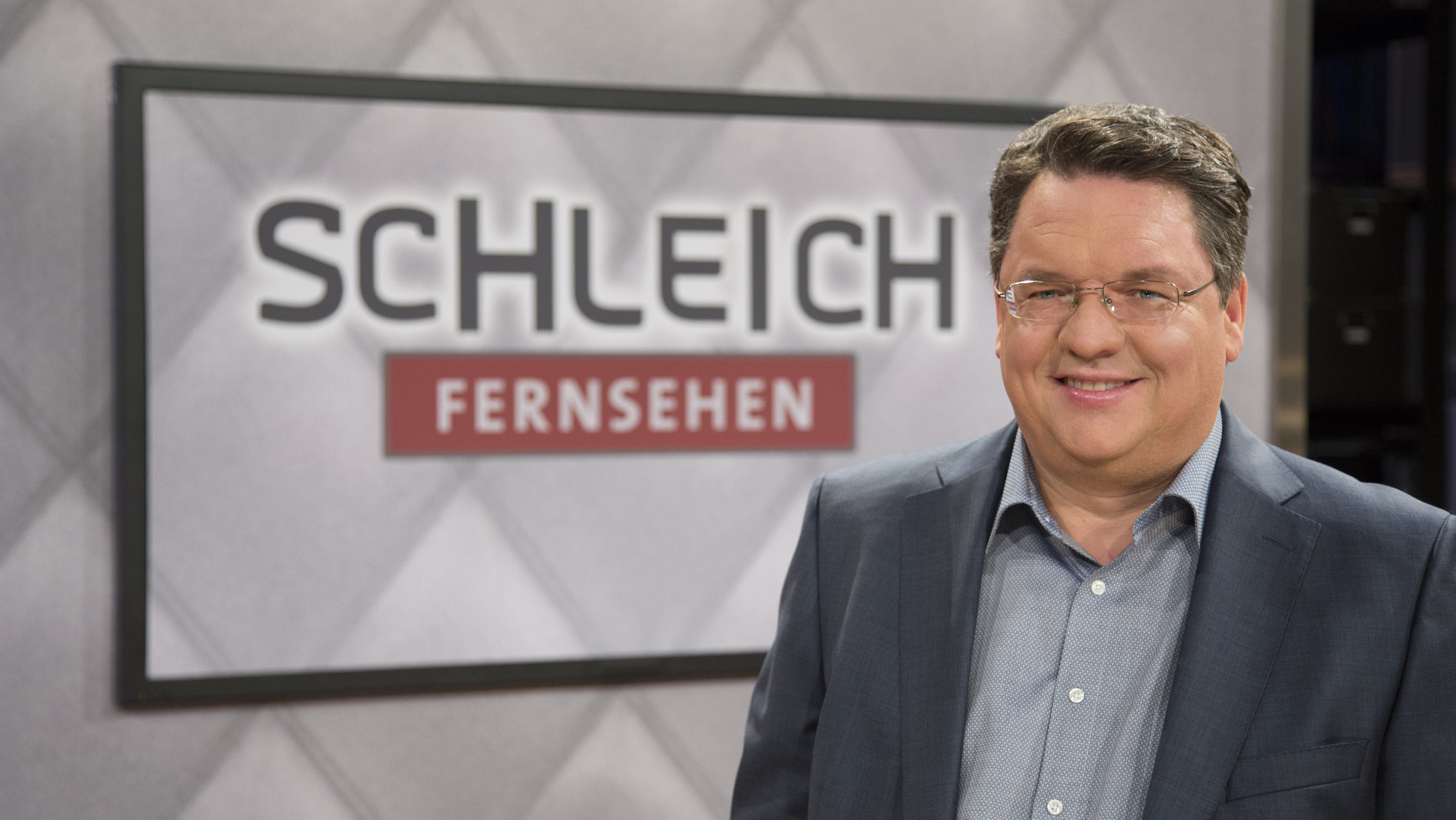 "SchleichFernsehen": Helmut Schleich (nur Oberkörper) vor einem Hintersetzer auf dem "Schleich Fernsehen" steht.