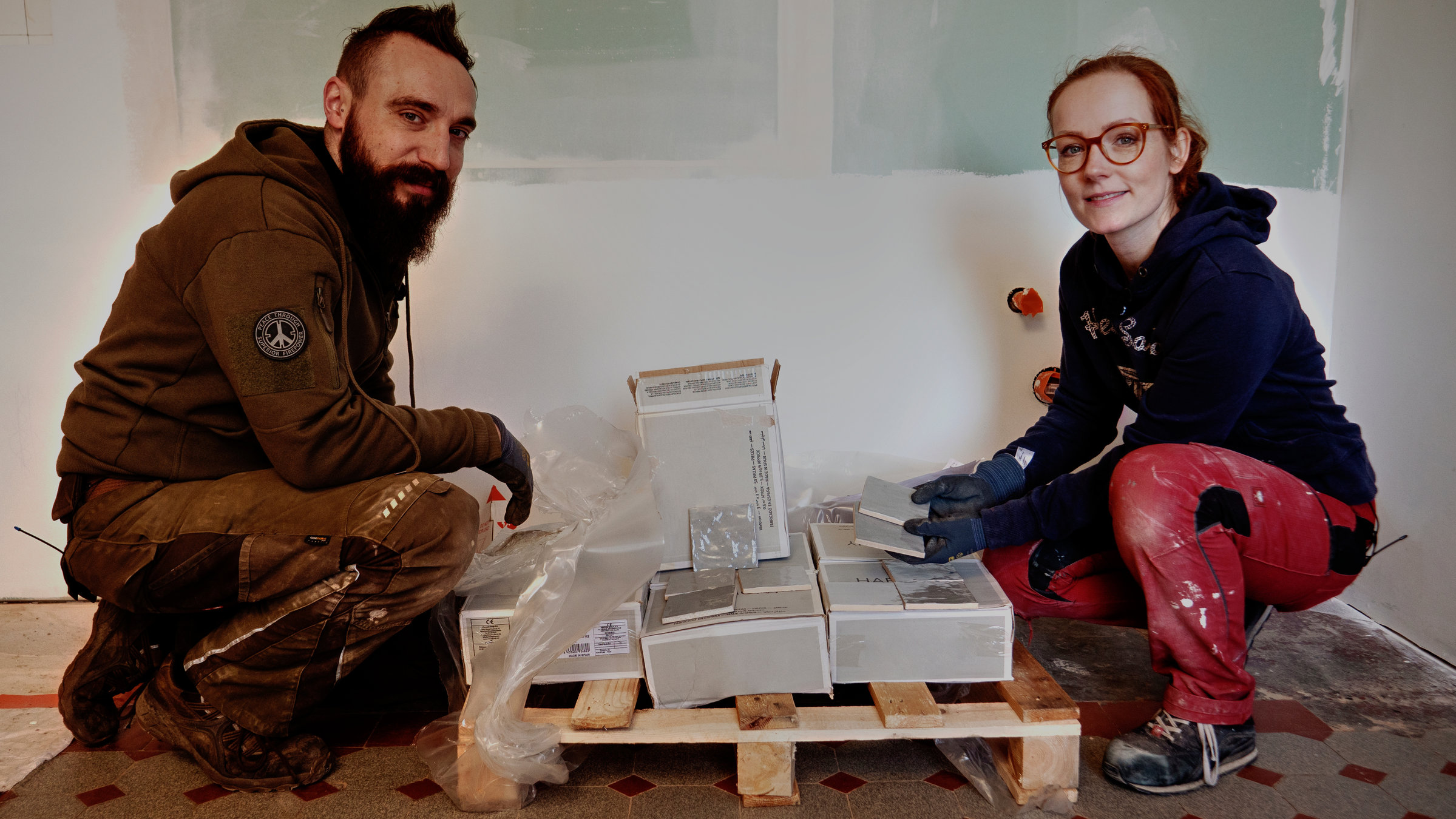 "ZDF.reportage: Wir bauen fertig - Der lange Weg zum Schnäppchenhaus": Falko und Janin tragen Arbeitskleidung. Zwischen den beiden steht auf dem Boden eine Palette mit Kartons, auf den Kartons liegen kleine quadratische Fliesen.