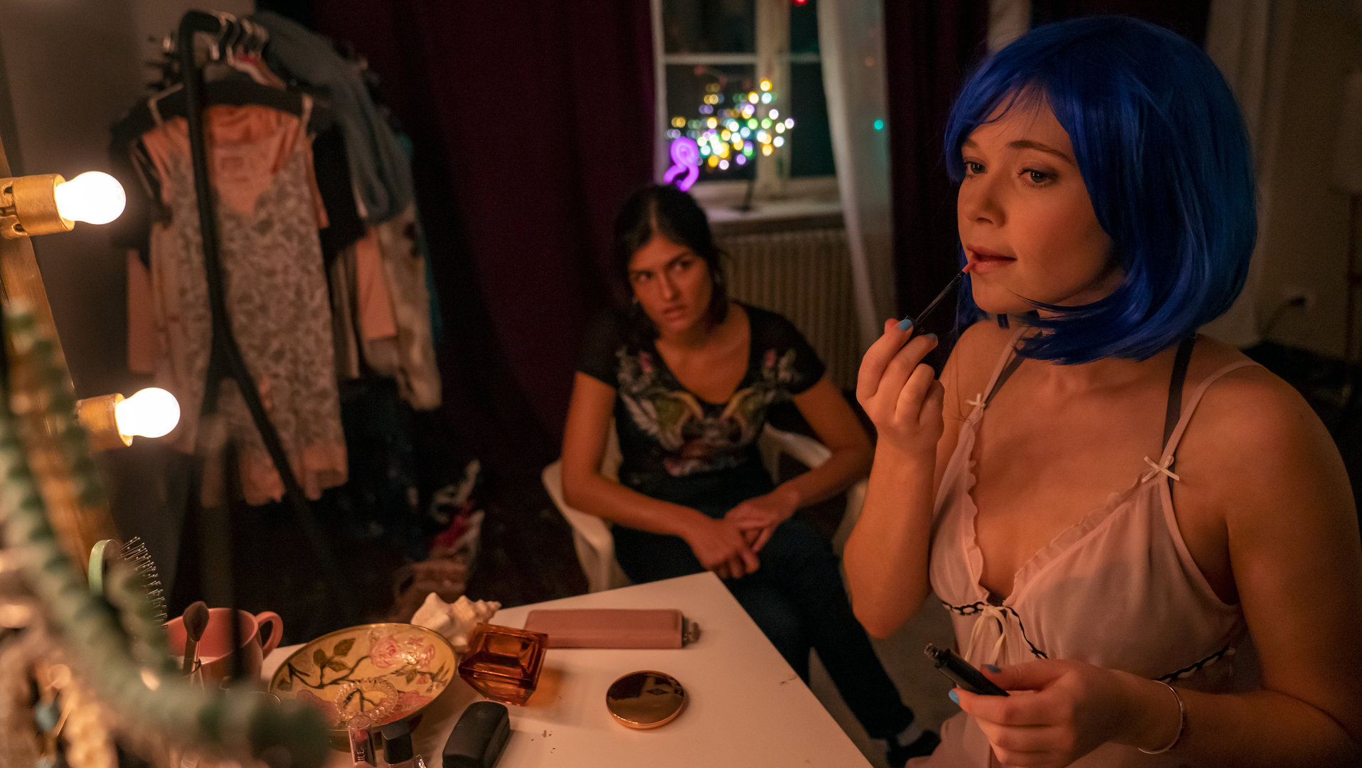 "Letzte Spur Berlin - Kleine Blume": Fiona (Farina Flebbe) und Lina (Julie Burkhardt) befinden sich in Fionas Schlafzimmer. Fiona sitzt mit blauer Perücke an ihrem Schminktisch und schminkt ihre Lippen mit einem Lipgloss. Lina sitzt auf einem Stuhl neben ihr und schaut sie fragend an.