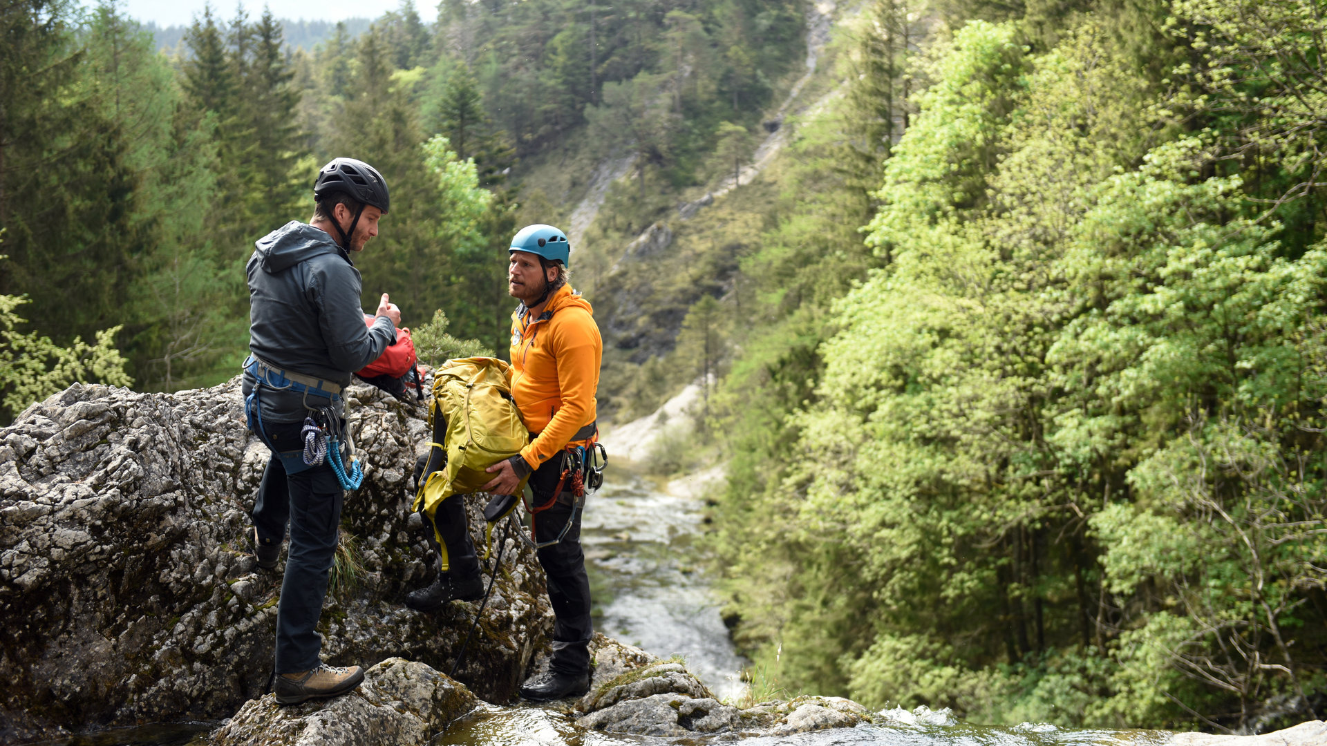 "Die Bergretter: Um jeden Preis": An einer Wasserfallklippe stehen sich Georg Haas (Leopold Hornung)und Bergretter Markus (Sebastian Ströbel) gegenüber. Sie tragen Helme und Kletterausrüstungen. Markus hält einen Rucksack in den Händen. Im Hintergrund ein bewaldeter Berghang.