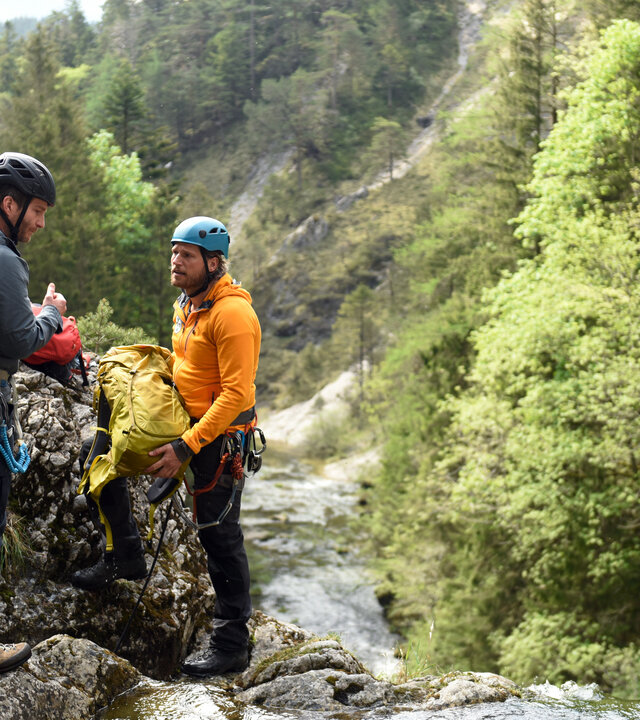 "Die Bergretter: Um jeden Preis": An einer Wasserfallklippe stehen sich Georg Haas (Leopold Hornung)und Bergretter Markus (Sebastian Ströbel) gegenüber. Sie tragen Helme und Kletterausrüstungen. Markus hält einen Rucksack in den Händen. Im Hintergrund ein bewaldeter Berghang.