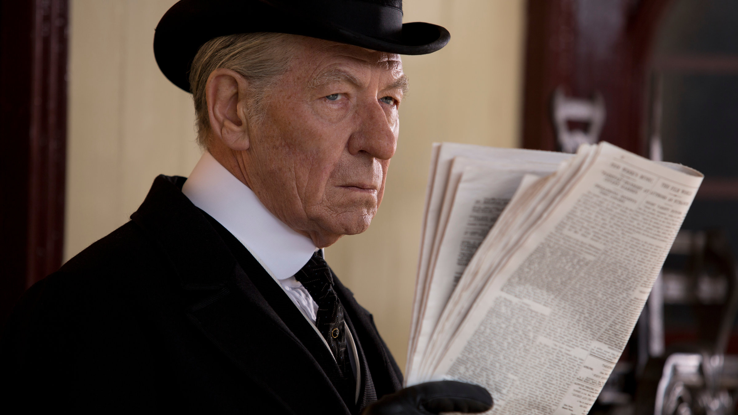 „Mr. Holmes“: Sherlock Holmes (Ian McKellen) in schwarzem Frack, mit Handschuhen und Zylinder blickt konzentriert von einer gefalteten Zeitung auf, die er in Händen hält.