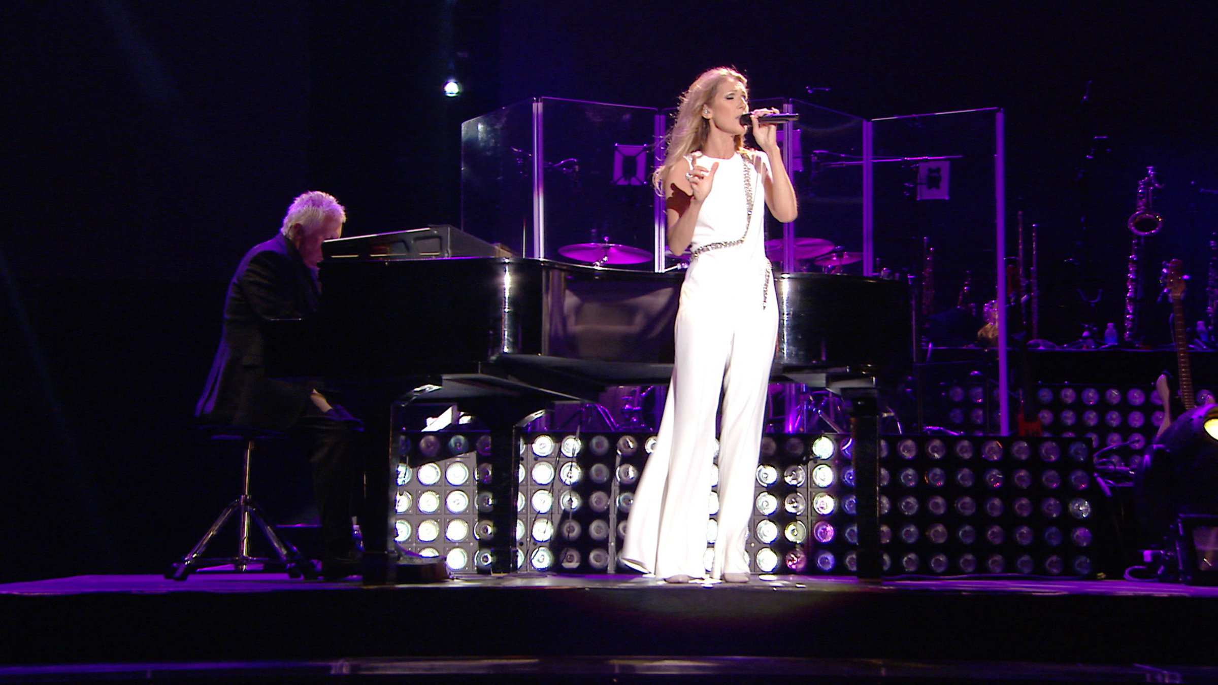 "Céline Dion: Une Seule Fois": Die Sängerin Céline Dion steht auf einer Bühne und singt. Gekleidet ist sie mit einem weißen ärmellosen Overall. Hinter ihr sitzt ein Mann an einem Flügel.