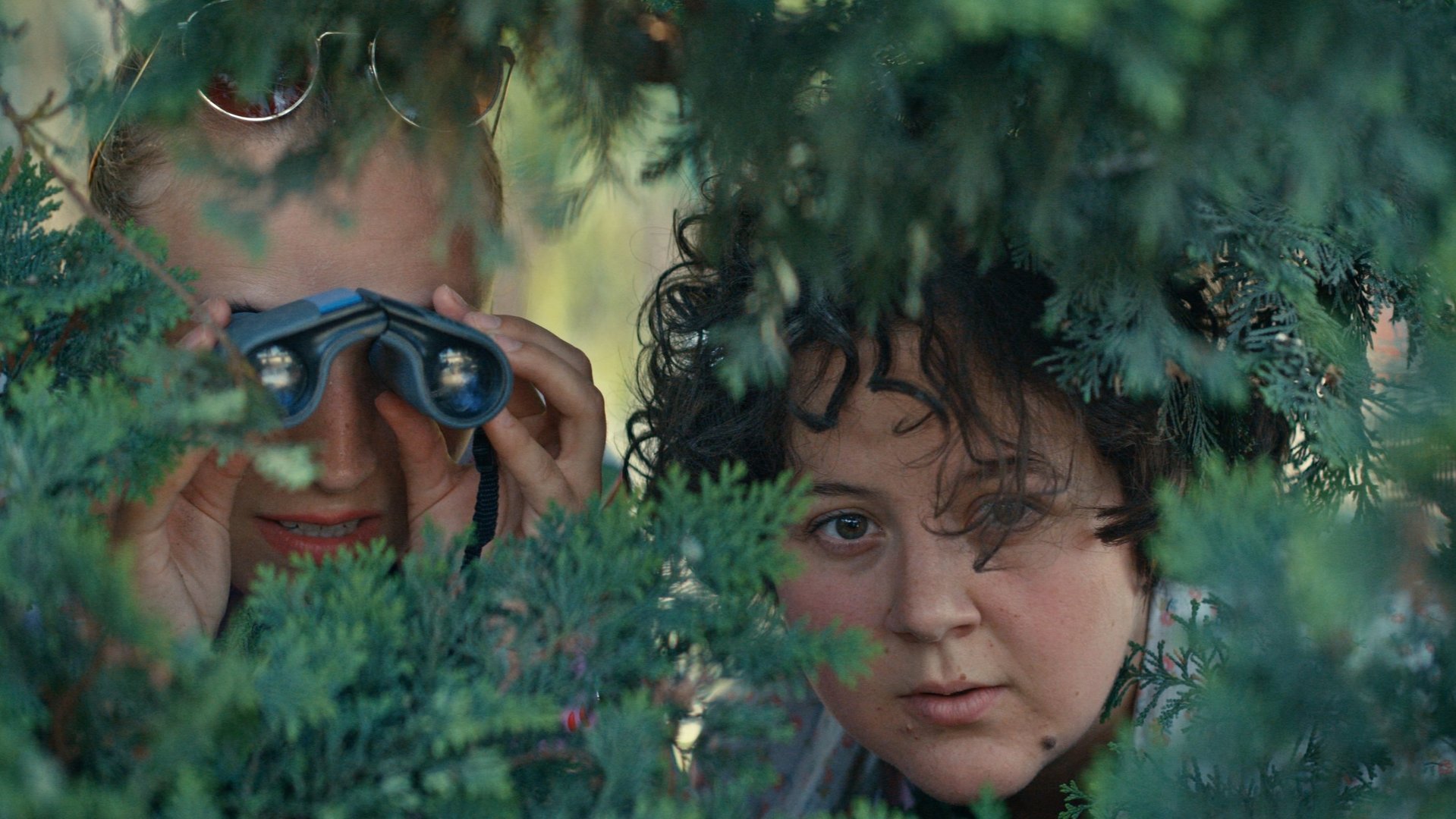 "Derby Girl - Putsch": Lola Bouvier (Chloé Jouannet) und Marie-Josée (Salomé Diénis Meulien) verstecken sich hinter einem Busch und blicken durch eine Lücke konzentriert in die Ferne. Lola hält sich dabei ein Fernglas vor ihre Augen.