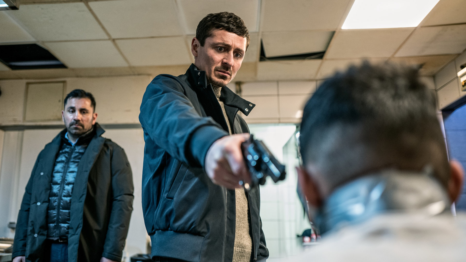 "Hidden Agenda II: Rache": Teddy (Alexej Manvelov) zielt mit einem Revolver auf den gefesselten Mahir (Wissam Rateb), der nur von hinten zu sehen ist. Isak (Mahmut Suvakci) hält sich im Hintergrund.