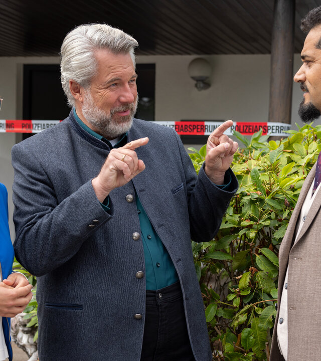 "Die Rosenheim-Cops - Ein brillantes Geschäft": Die Kommissare Beck (Michaela Weingartner) und Stadler (Dieter Fischer) stehen vor der Hecke vor einem Wohnhaus und sprechen mit Luis Spohr (Patrick Pinheiro), der ihnen gegenübersteht. Stadler gestikuliert, während er Spohr ansieht.