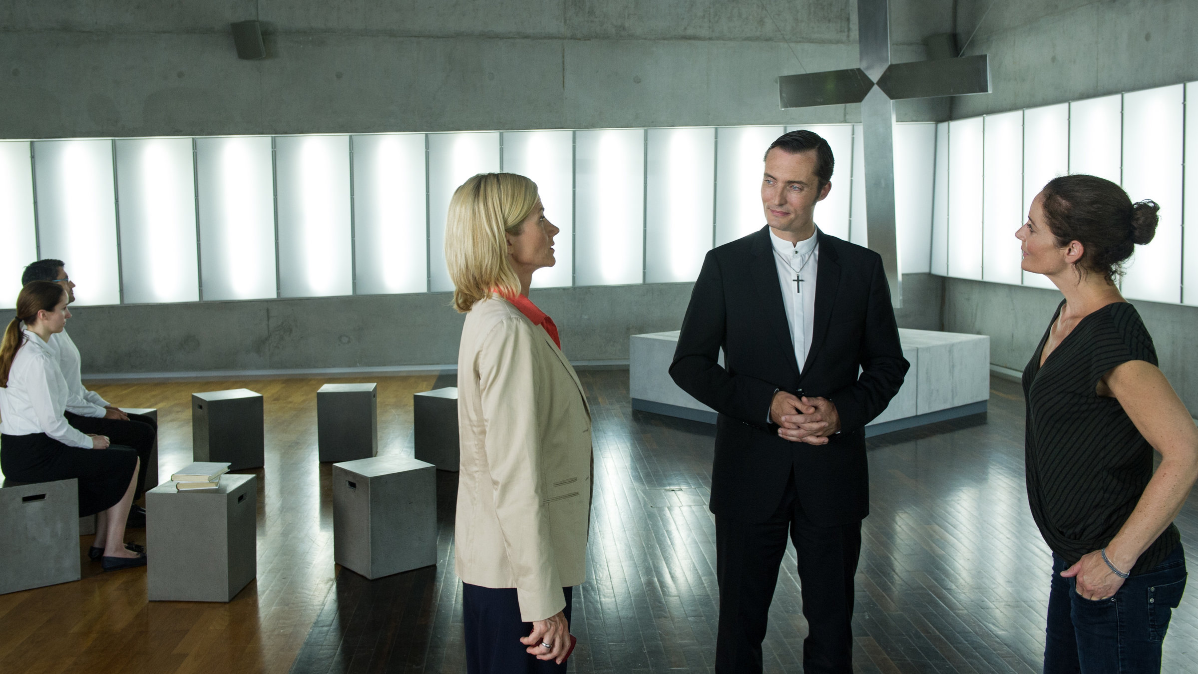 "SOKO Stuttgart - Im Namen Gottes": Martina Seiffert (Astrid M. Fünderich) und Selma Kirsch (Yve Burbach) sprechen mit Niklas Reich (Marco Di Sapia), der zwischen ihnen steht. Links seitlich sitzen zwei Gemeindemitglieder (Komparsen) und warten geduldig im Gemeindesaal der Freikirche. Im Hintergrund ist ein großes chromfarbenes Kreuz zu sehen.