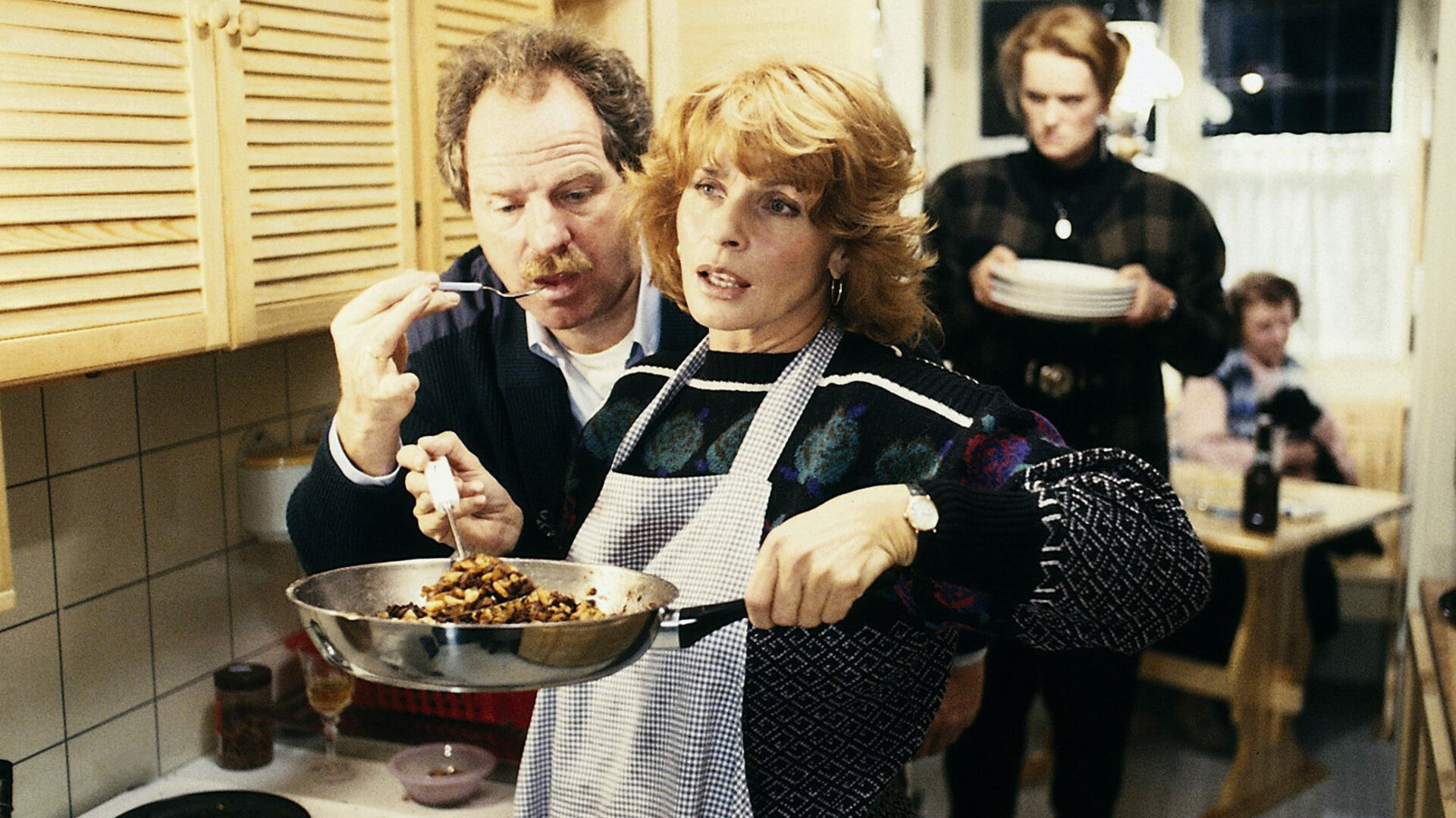 "Die schnelle Gerdi - Gerdi gründet eine Familie": Gerdi (Senta Berger) kocht in einer Küche und hält eine Pfanne hoch, während Rudi (Friedrich von Thun) dicht hinter ihr steht und ihr etwas kostet. Im Hintergrund trägt Jennifer (Elena Rublack) Teller in der Hand.