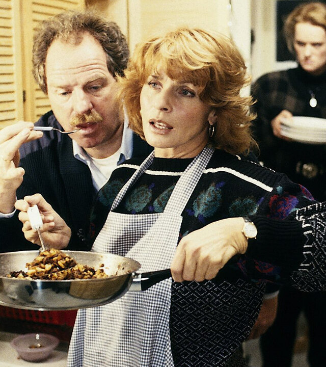 "Die schnelle Gerdi - Gerdi gründet eine Familie": Gerdi (Senta Berger) kocht in einer Küche und hält eine Pfanne hoch, während Rudi (Friedrich von Thun) dicht hinter ihr steht und ihr etwas kostet. Im Hintergrund trägt Jennifer (Elena Rublack) Teller in der Hand.
