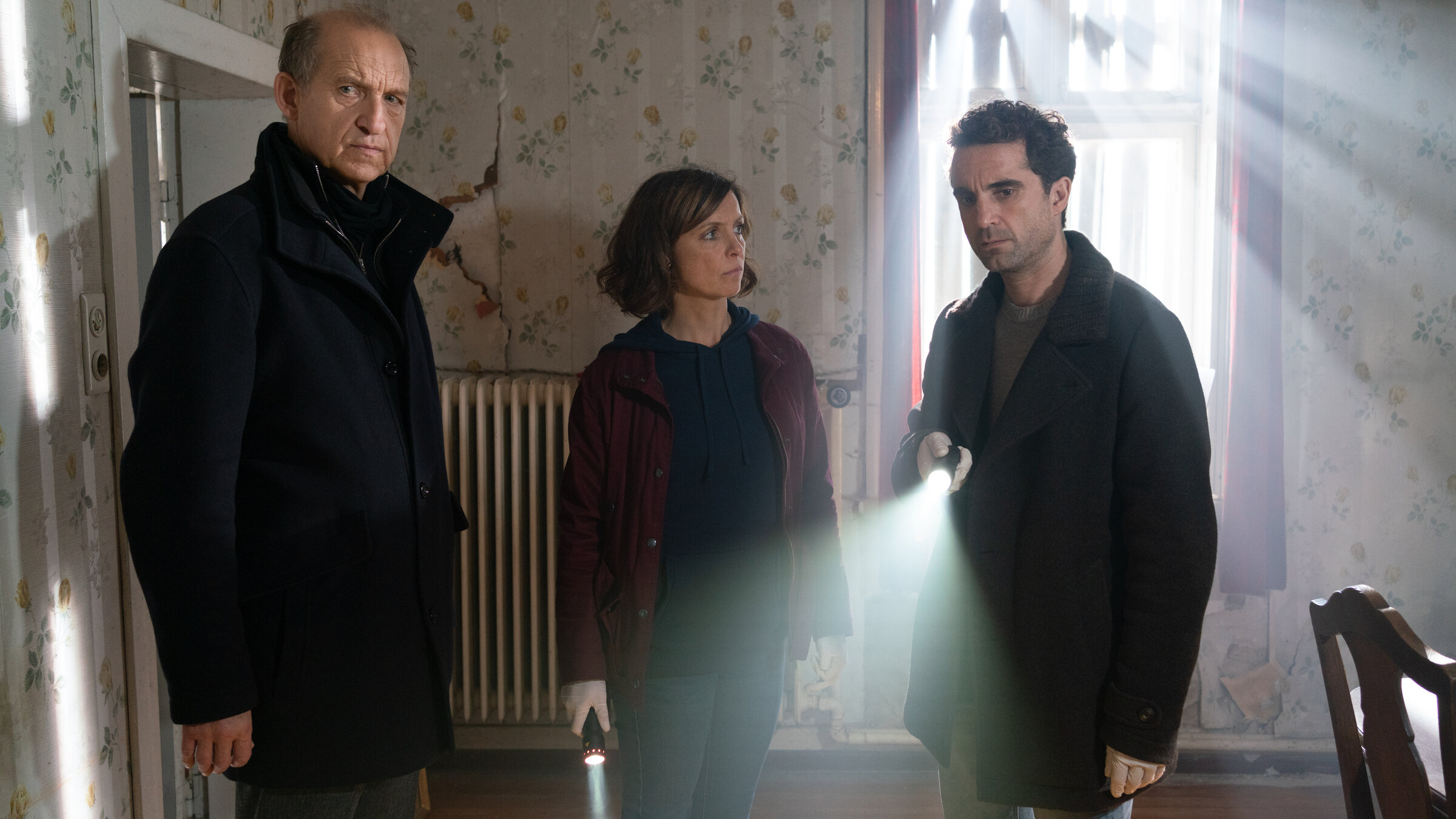 "Nord Nord Mord - Sievers und das Geisterhaus": Carl Sievers (Peter Heinrich Brix) steht neben Ina Behrendsen (Julia Brendler) und Hinnerk Feldmann (Oliver Wnuk) in einem Raum mit heruntergekommener Tapete mit Blumenmuster. Hinter Hinnerk befindet sich ein mit Brettern zugenageltes Fenster, durch das Licht einfällt. Ina und Hinnerk halten jeweils eine Taschenlampe in den Händen. Ina sieht Hinnerk ernst an. Dieser blickt, so wie Sievers, nachdenklich nach vorne.