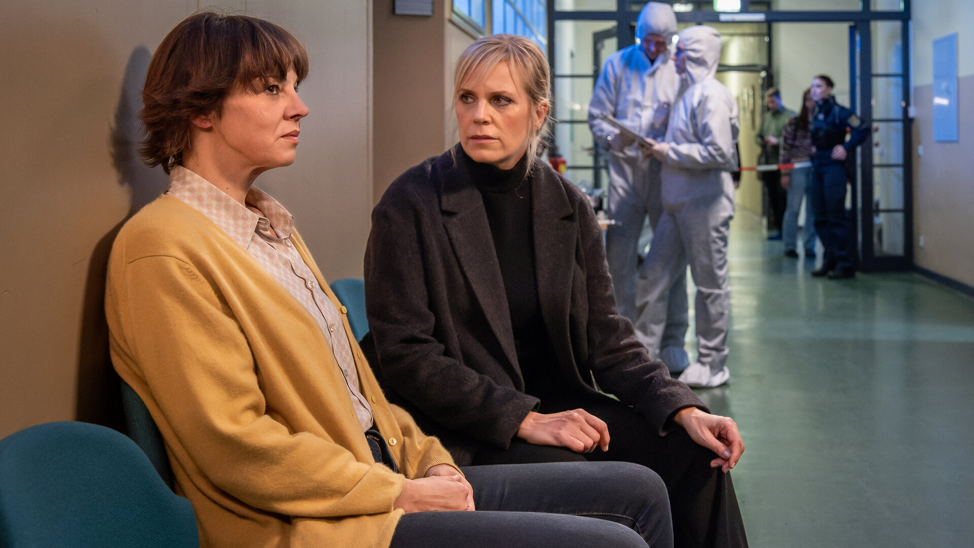 "SOKO Leipzig - Unter Druck": Kathrin Schwake (Doreen Fietz) und Ina Zimmermann (Melanie Marschke) sitzen nebeneinander auf dem Gang der Schule. Die Spurensicherung geht im Hintergrund ihrer Arbeit nach (Komparsen).
