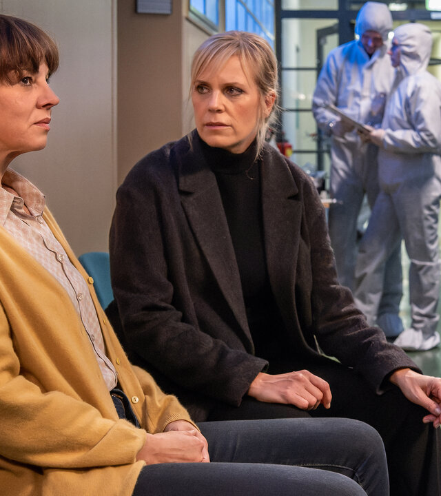 "SOKO Leipzig - Unter Druck": Kathrin Schwake (Doreen Fietz) und Ina Zimmermann (Melanie Marschke) sitzen nebeneinander auf dem Gang der Schule. Die Spurensicherung geht im Hintergrund ihrer Arbeit nach (Komparsen).