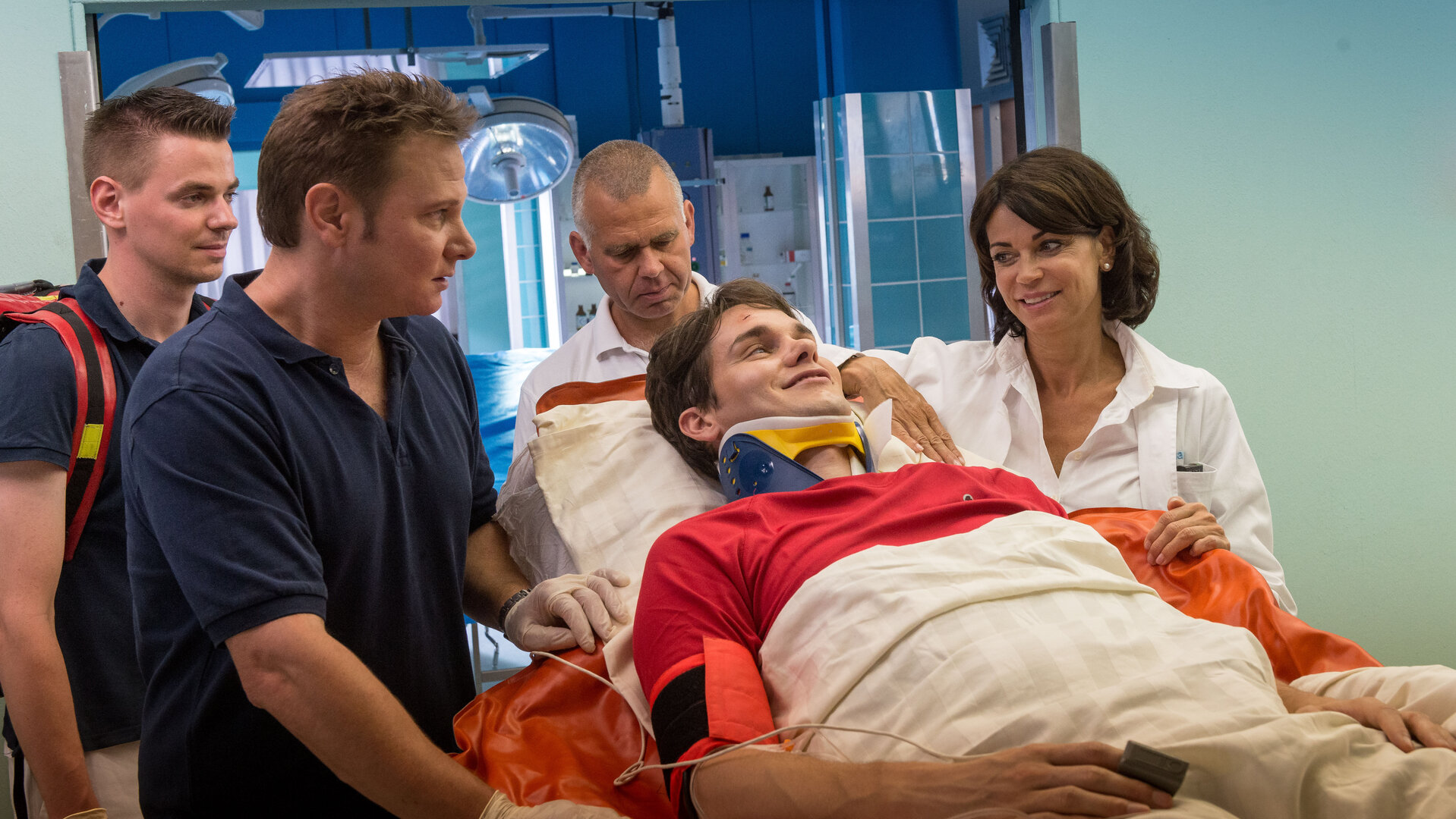 "Notruf Hafenkante - Sport ist Mord": Josh Baier (Daniel Fünffrock) wird in das Elbkrankenhaus eingeliefert. Er liegt auf eine Krankentrage. An den beiden Seiten der Trage stehen Notarzt Dr. Haase (Fabian Harloff) und Dr. Jasmin Jonas (Gerit Kling). Hinter der Trage stehen zwei weitere Männer des Pflegepersonals.