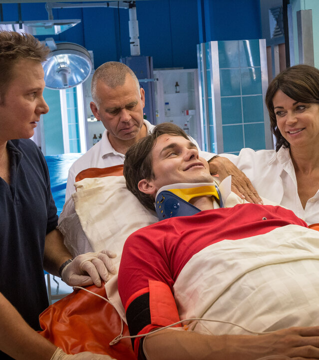 "Notruf Hafenkante - Sport ist Mord": Josh Baier (Daniel Fünffrock) wird in das Elbkrankenhaus eingeliefert. Er liegt auf eine Krankentrage. An den beiden Seiten der Trage stehen Notarzt Dr. Haase (Fabian Harloff) und Dr. Jasmin Jonas (Gerit Kling). Hinter der Trage stehen zwei weitere Männer des Pflegepersonals.