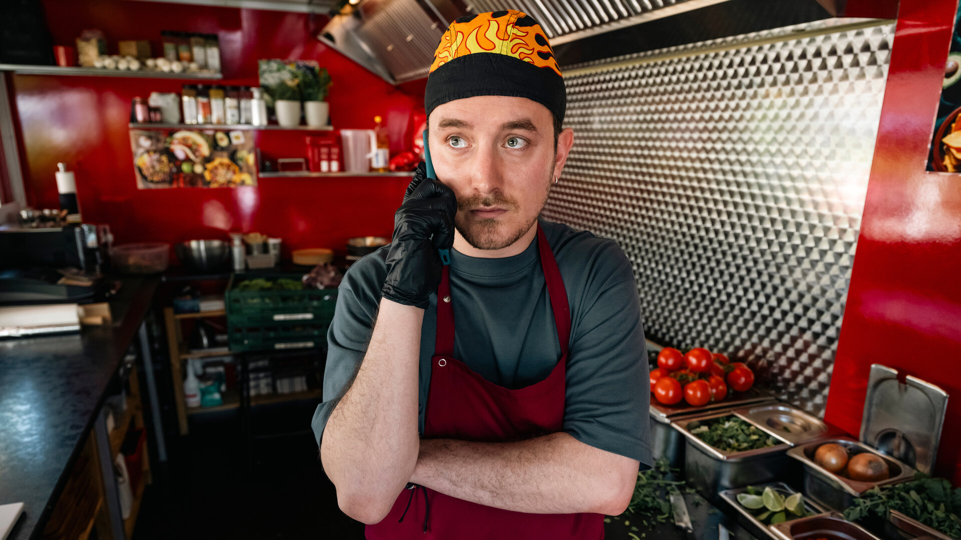 "SOKO Köln - Kantinentod": Chris Palewski (David Hugo Schmitz) steht in einem rot lackiertem Foodtruck und hält sich mit ernster Miene sein Handy ans Ohr. Er trägt schwarze Gummihandschuhe, eine rote Schürze und eine schwarze Mütze, die mit Flammen bedruckt ist. Neben ihm steht diverses Gemüse in silbernen Behältern.