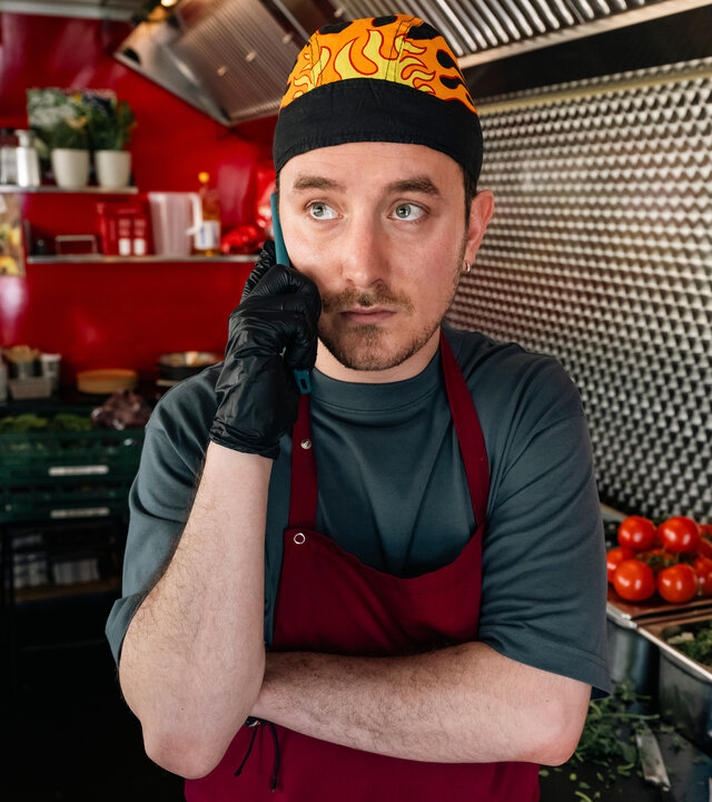 "SOKO Köln - Kantinentod": Chris Palewski (David Hugo Schmitz) steht in einem rot lackiertem Foodtruck und hält sich mit ernster Miene sein Handy ans Ohr. Er trägt schwarze Gummihandschuhe, eine rote Schürze und eine schwarze Mütze, die mit Flammen bedruckt ist. Neben ihm steht diverses Gemüse in silbernen Behältern.