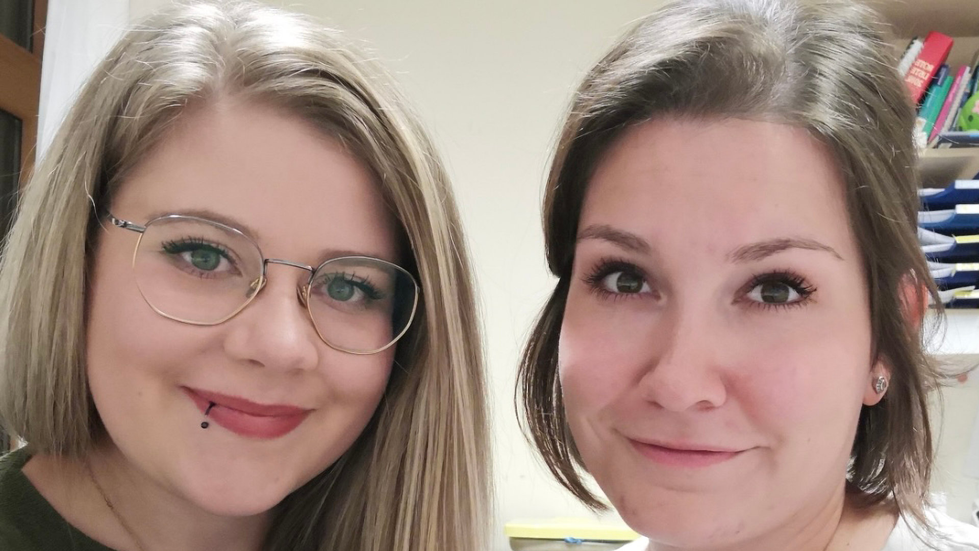 "Leben ist mehr! - Zum Karfreitag 2021": Porträtaufnahme von zwei jungen Frauen. Links im Bild Michaela B. mit blonden langen Haare, Brillenträgerin. Merkmal Piercingschmuck an der rechten Lippe. Sie trägt ein olivfarbenes Oberteil. Rechts im Bild Sara L. Die junge Frau hat braunes schulterlanges Haar. Sie trägt ein weißes Oberteil. Im Hintergrund ein Bücherregal.