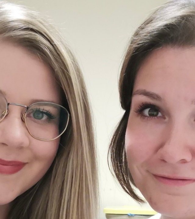 "Leben ist mehr! - Zum Karfreitag 2021": Porträtaufnahme von zwei jungen Frauen. Links im Bild Michaela B. mit blonden langen Haare, Brillenträgerin. Merkmal Piercingschmuck an der rechten Lippe. Sie trägt ein olivfarbenes Oberteil. Rechts im Bild Sara L. Die junge Frau hat braunes schulterlanges Haar. Sie trägt ein weißes Oberteil. Im Hintergrund ein Bücherregal.