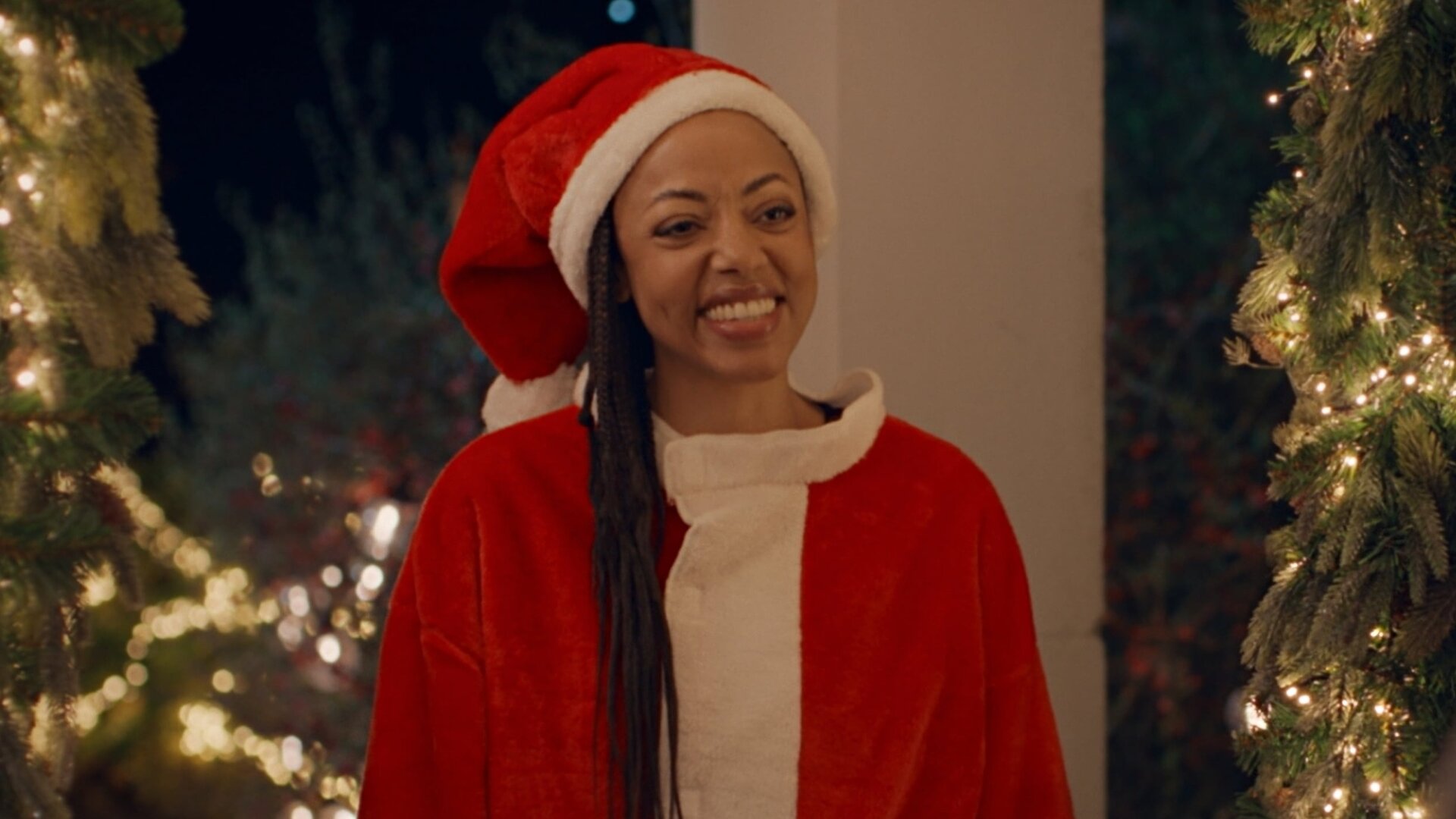 "Single Bells - 12 Dates till Christmas: Alex": Alex (Sandrine Van Handenhoven), eine strahlend lächelnde junge Frau mit langen, schwarzen Haaren, steht in einem Nikolauskostüm vor einer Haustür. Im Hintergrund leuchtet Weihnachtsschmuck.