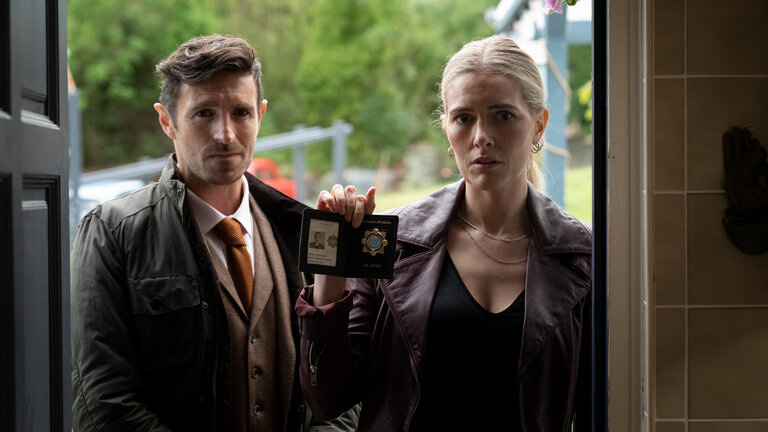 "Borderline – Grenzenlose Verbrechen - Bis dass der Tod sie scheidet": DCI Philip Boyd (Eoin Macken) und DI Aoife Regan (Amy De Bhrún) stehen nebeneinander vor einer geöffneten Haustür. Regan hält ihren Dienstausweis hoch.