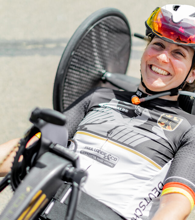 "einfach Mensch - Annika Zeyen-Giles: Paralympics 2024 (3)": Annika Zeyen-Giles liegt in ihrem Wettkampf-Handbike und lacht in die Kamera.