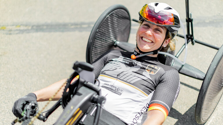 "einfach Mensch - Annika Zeyen-Giles: Paralympics 2024 (3)": Annika Zeyen-Giles liegt in ihrem Wettkampf-Handbike und lacht in die Kamera.