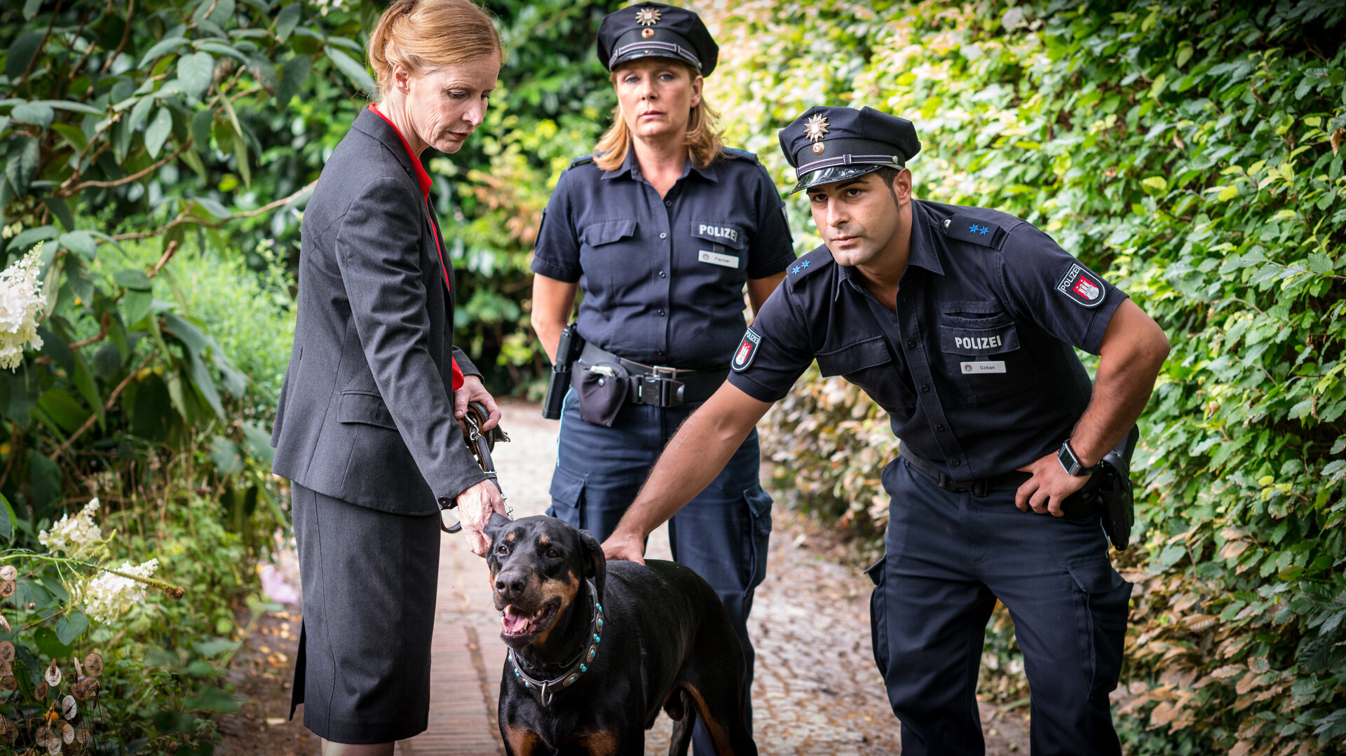 "Notruf Hafenkante - Hund und Katze": Petra Wolter (Konstanze Proebster) steht in ihrem Garten mit ihrem Hund an der Leine. Der Polizeibeamte Tarik Coban (Serhat Cokgezen) streichelt den Hund. Seine Kollegin Claudia Fischer (Janette Rauch) steht, die Arme auf dem Rücken verschränkt, zwischen den beiden.
