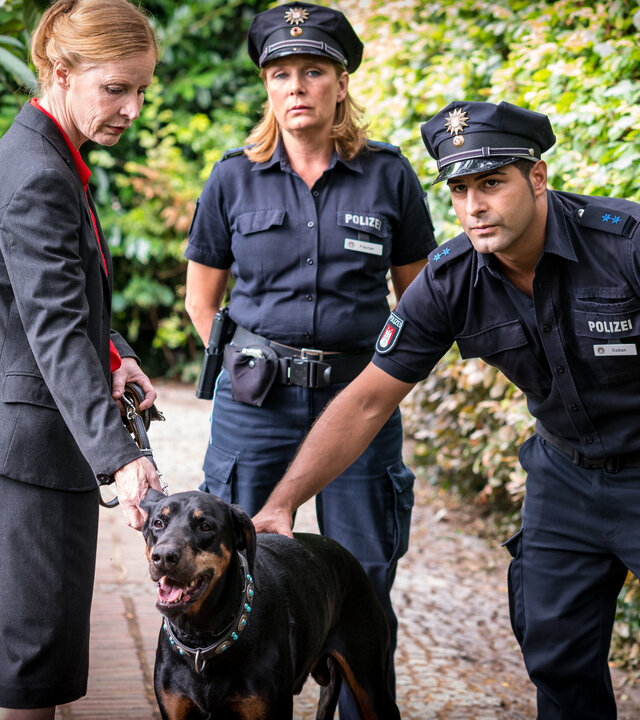 "Notruf Hafenkante - Hund und Katze": Petra Wolter (Konstanze Proebster) steht in ihrem Garten mit ihrem Hund an der Leine. Der Polizeibeamte Tarik Coban (Serhat Cokgezen) streichelt den Hund. Seine Kollegin Claudia Fischer (Janette Rauch) steht, die Arme auf dem Rücken verschränkt, zwischen den beiden.