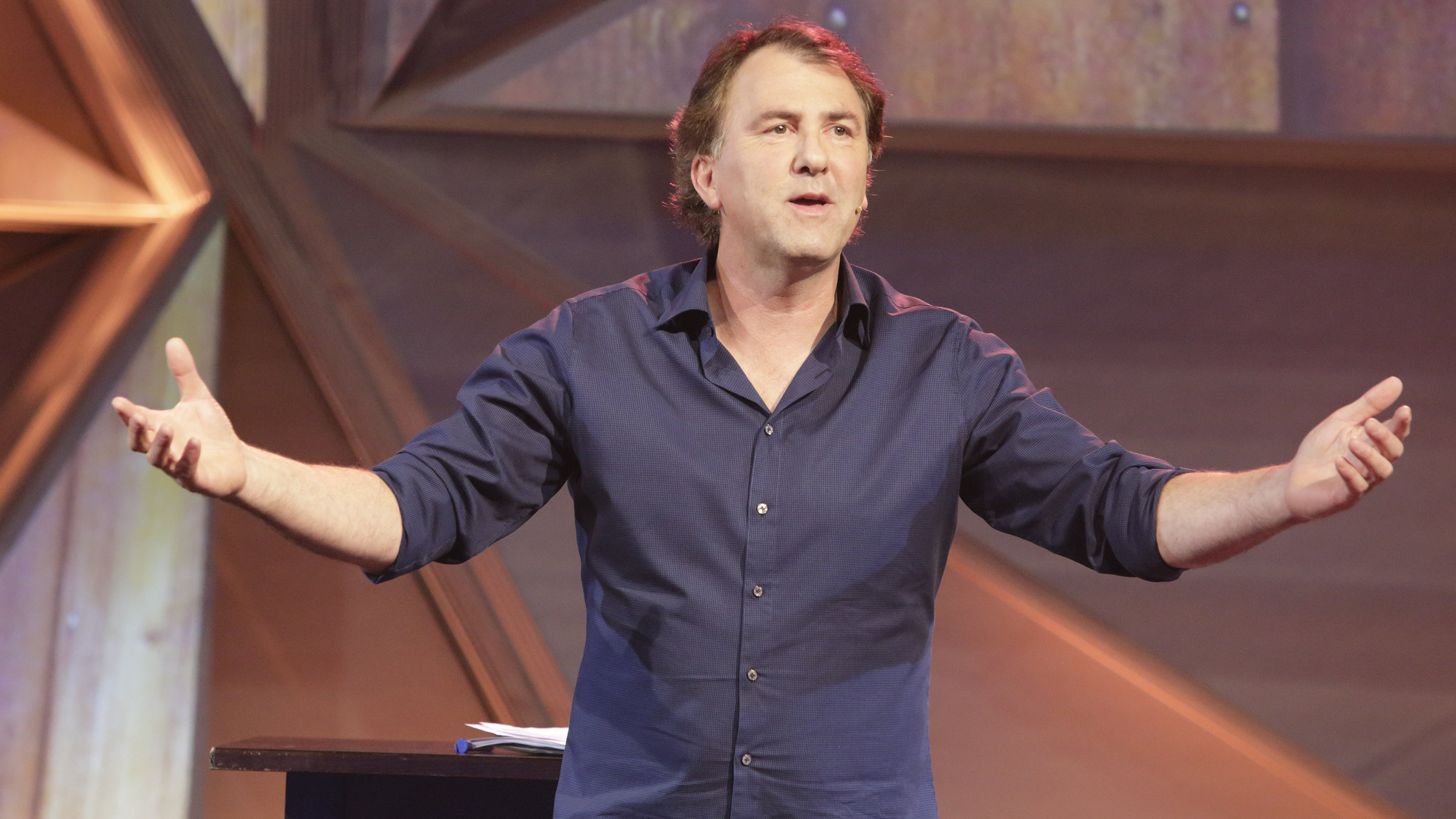 "Stefan Waghubinger: Außergewöhnliche Belastungen": Stefan Waghubinger trägt ein dunkelblaues Hemd und eine graue Hose. Er steht auf der Bühne vor einem Tisch auf dem mehrere Zettel liegen.