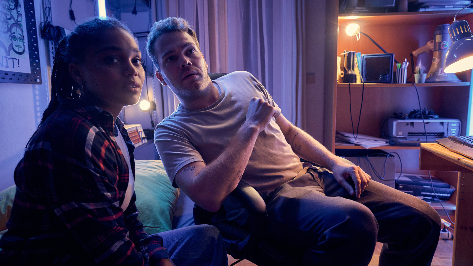 "Like a Loser - Bierernst": Lea (Sira Anna Faal) und Julian (Ben Münchow) sitzen im Kinderzimmer von Ernst (Diyar Ilhan) – Lea auf dem Bett, Julian auf dem Schreibtischstuhl. Das Zimmer ist nur spärlich beleuchtet. Beide blicken überrascht in dieselbe Richtung.