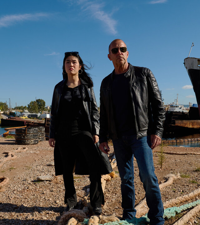 "Yvonne und der Tod": Ingo Thiel (Heino Ferch) und Lee Sooyoung (Kotbong Yang) stehen zwischen Tauen, die Schiffe im Hintergrund befestigen. Beide haben den Blick kritisch in die Ferne gerichtet. Ingo Thiel trägt eine Sonnenbrille.