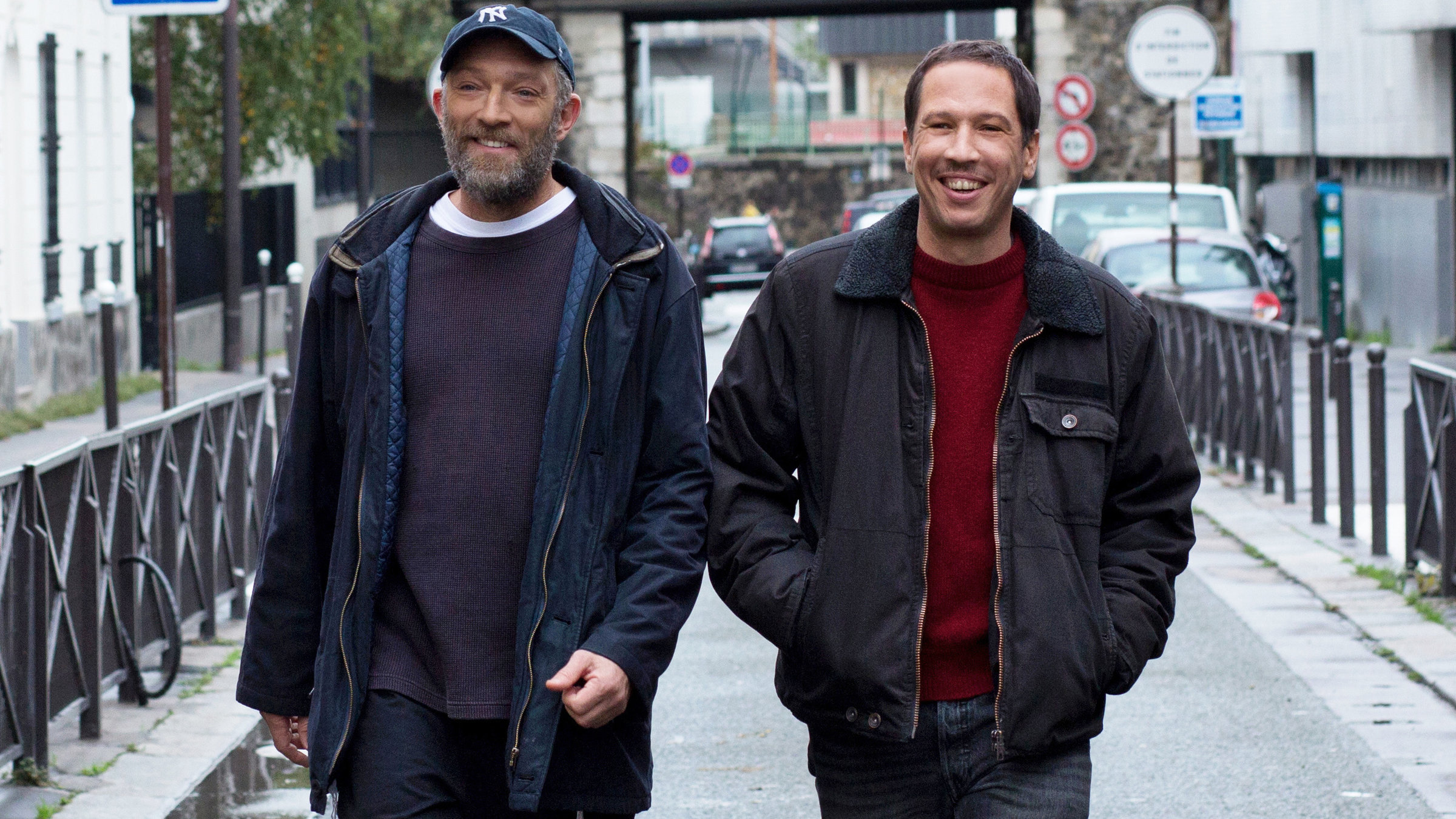 "Alles außer gewöhnlich": Bruno, ein Mann mittleren Alters mit grauem Bart, Schildmütze und dunkelblauem Parka geht mit Malik, ebenfalls mittleren Alters mit Schnurrbart, rotem Pulli und dunklem Anorak über eine Straße in Paris. Beide lächeln, Malik hat die Hände in den Jackentaschen.