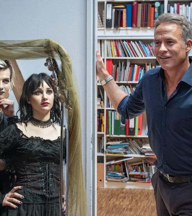 "SOKO Leipzig - Schwarz ist alle Farben": Jojo Keller (Karl Seibt) und Lotte Trautzschke (Carlotta Bähre) sind über einen Spiegel in Gothic-Klamotten zu sehen. Beide lächeln Jan Maybach (Marco Girnth) herausfordernd an, welcher in der Zimmertür steht und leicht überfordert lächelt.