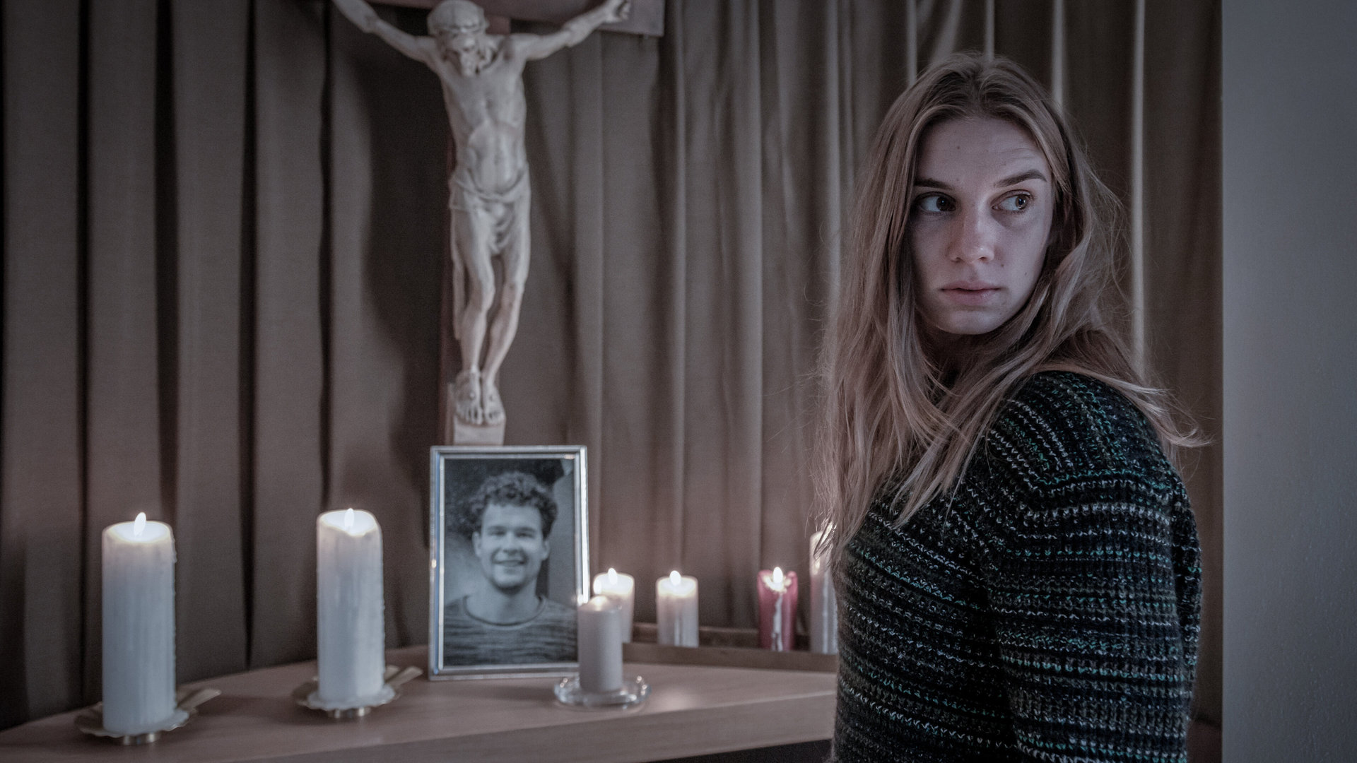 "SOKO Linz - Lazarus": Clara (Maya Unger) steht vor dem Altar ihres Bruders Philipps, auf dem ein Porträt von ihm und leuchtende Kerzen stehen. Über dem Altar hängt eine Statue von Jesus am Kreuz.