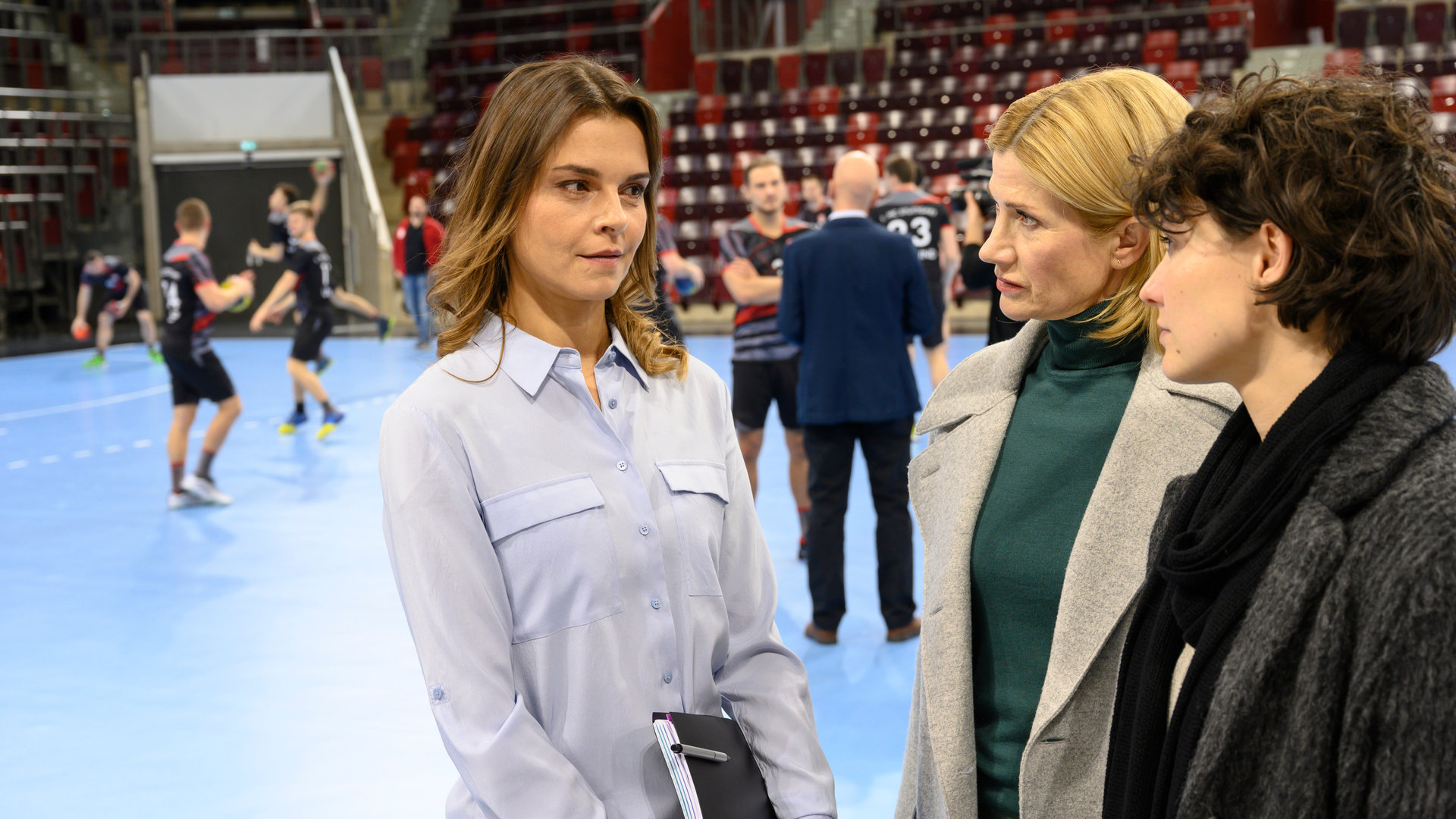 "SOKO Stuttgart – Abstiegskampf": Martina Seiffert (Astrid M. Fünderich) steht mit ihrer Kollegin Nele Becker (Nina Siewert) auf dem blauen Spielfeld einer Handballarena und befragt Annika Brett (Susan Hoecke), die Pressesprecherin des "1. HC Stuttgart". Im Hintergrund sind Handballspieler (Komparsen) beim Training zu sehen. Die Zuschauerränge sind unbesetzt. Der Star der Mannschaft, Florian Mayer (Daniel Axt) unterhält sich mit einem Mann im dunklen Anzug.