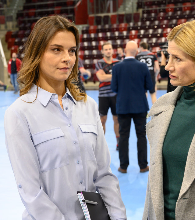 "SOKO Stuttgart – Abstiegskampf": Martina Seiffert (Astrid M. Fünderich) steht mit ihrer Kollegin Nele Becker (Nina Siewert) auf dem blauen Spielfeld einer Handballarena und befragt Annika Brett (Susan Hoecke), die Pressesprecherin des "1. HC Stuttgart". Im Hintergrund sind Handballspieler (Komparsen) beim Training zu sehen. Die Zuschauerränge sind unbesetzt. Der Star der Mannschaft, Florian Mayer (Daniel Axt) unterhält sich mit einem Mann im dunklen Anzug.