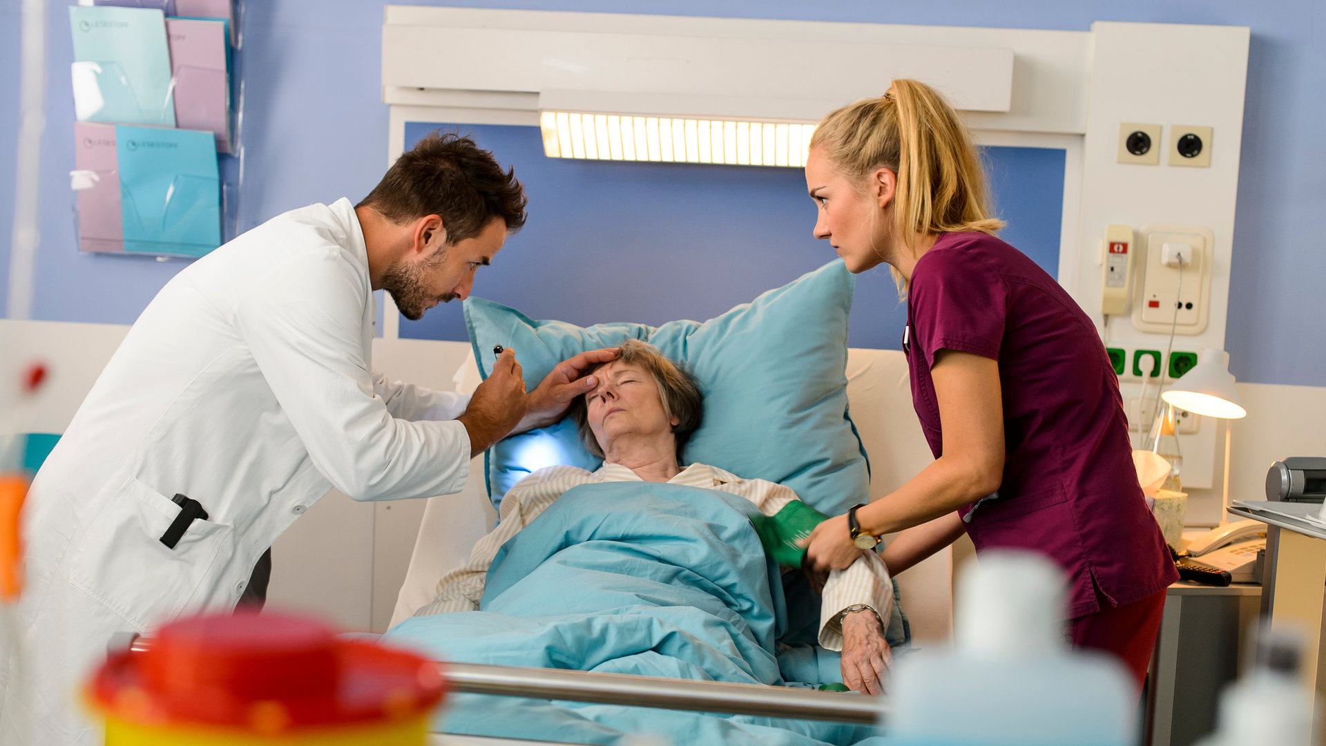 "Bettys Diagnose - Auf Liebe und Tod": Dr. Frank Stern (Max Alberti) und Betty Weiss (Annina Hellenthal) stehen auf beiden Seiten des Betts von Ilse Stern (Monika Lennartz) zu retten, die bewusstlos im Bett liegt. Dr. Stern hat eine Hand an ihrer Stirn und in der anderen eine Stableuchte, Betty legt ihr am anderen Arm ein Blutdruckmessgerät an.