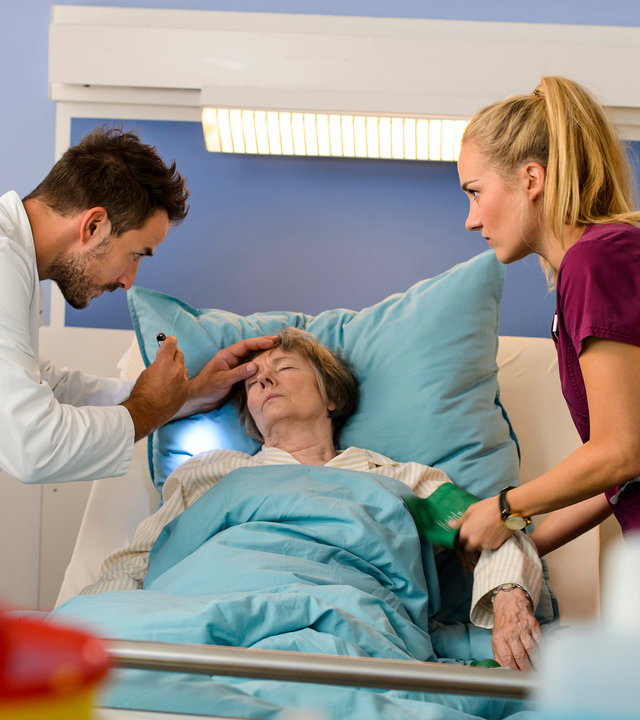 "Bettys Diagnose - Auf Liebe und Tod": Dr. Frank Stern (Max Alberti) und Betty Weiss (Annina Hellenthal) stehen auf beiden Seiten des Betts von Ilse Stern (Monika Lennartz) zu retten, die bewusstlos im Bett liegt. Dr. Stern hat eine Hand an ihrer Stirn und in der anderen eine Stableuchte, Betty legt ihr am anderen Arm ein Blutdruckmessgerät an.