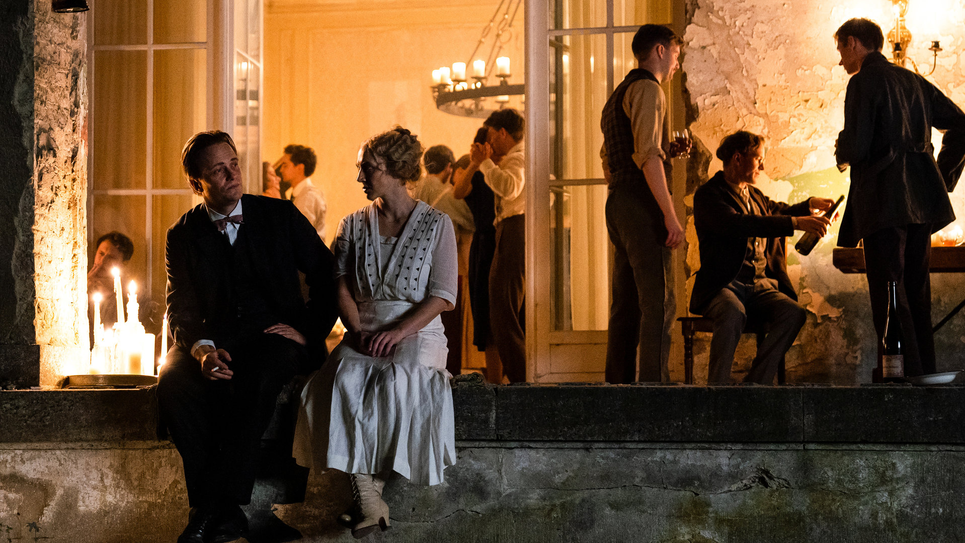 "Die Neue Zeit - 1. Nach dem Krieg": Walter Gropius (August Diehl) und Dörte Helm (Anna Maria Mühe) sitzen auf der Mauerumrandung einer Veranda und unterhalten sich. Im Hintergrund findet eine Party mit zahlreichen Kommilitonen statt. Kerzen verbreiten eine angenehme Atmosphäre.