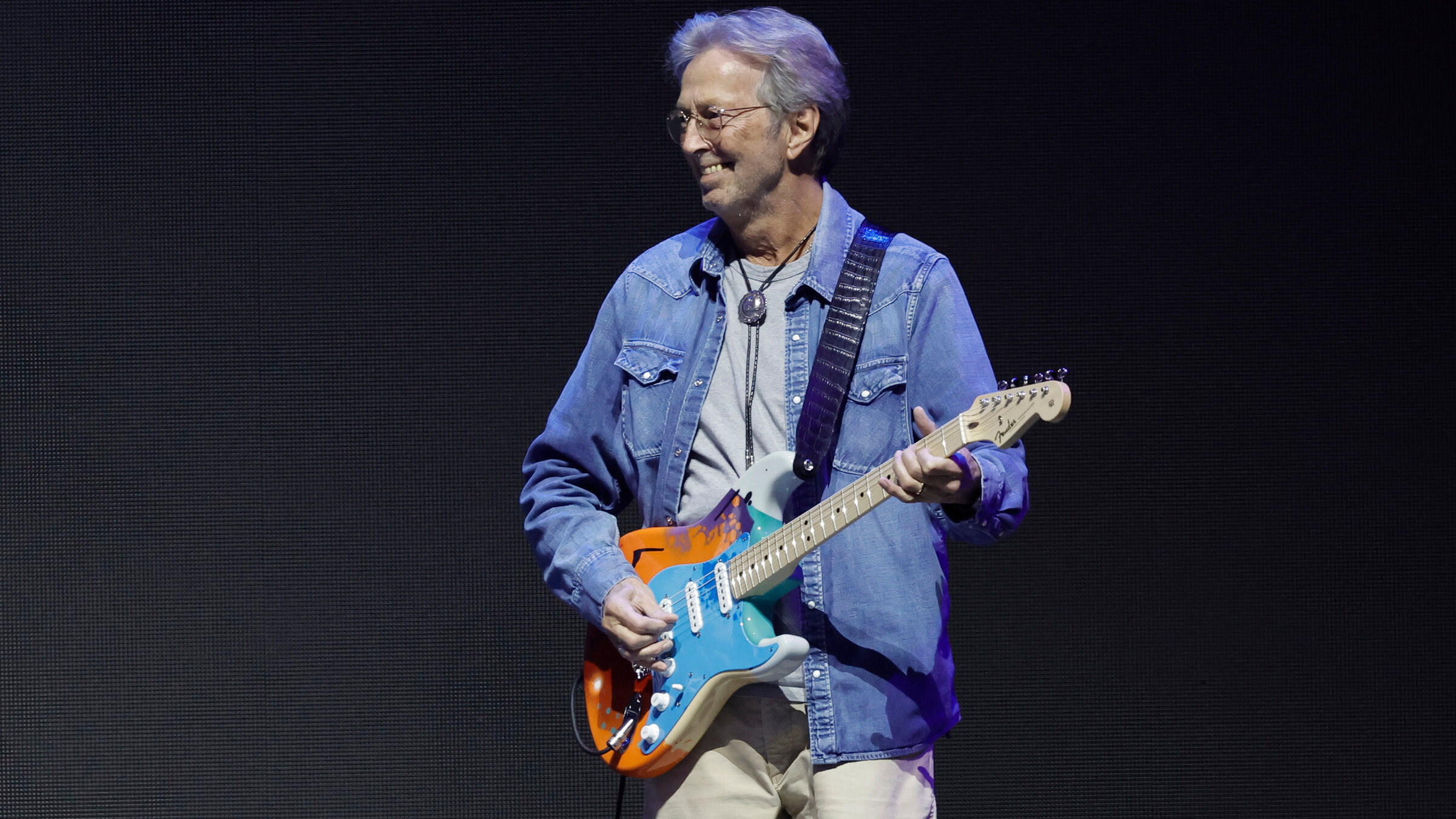 "Eric Clapton: Crossroads Guitar Festival 2023": Der Musiker Eric Clapton steht vor einem schwarzen Hintergrund. Er hält eine Gitarre in der Hand und blickt lächelnd zur Seite. Gekleidet ist er mit einem Jeanshemd und einer beigen Hose.