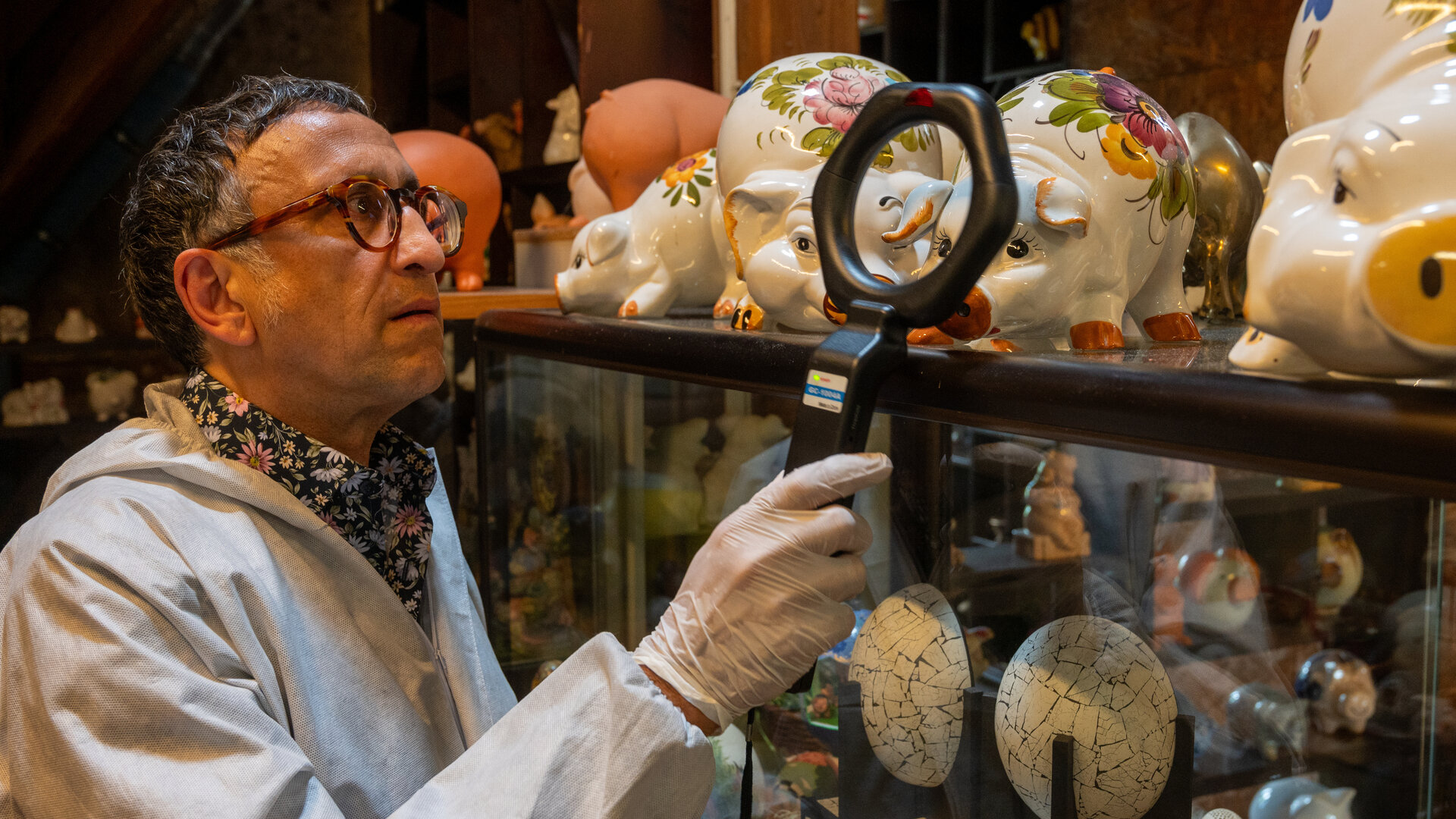 "SOKO Stuttgart - Perlen vor die Säue": Der Leiter der KTU Jan Arnaud (Mike Zaka Sommerfeldt) hält, gekleidet in Schutzanzug und mit Einweghandschuhen, einen Detektor an ein Porzellanschwein, das neben anderen Exponaten auf einer Vitrine steht.