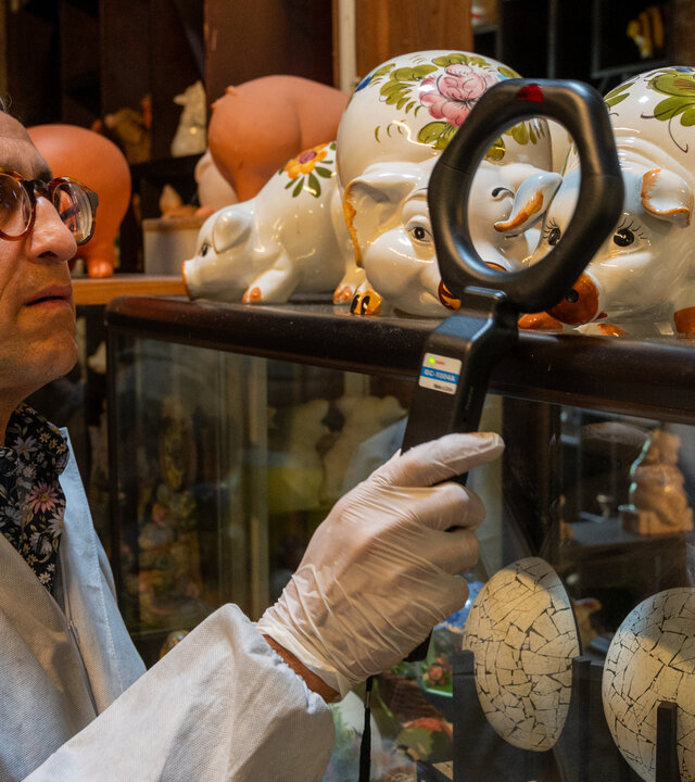 "SOKO Stuttgart - Perlen vor die Säue": Der Leiter der KTU Jan Arnaud (Mike Zaka Sommerfeldt) hält, gekleidet in Schutzanzug und mit Einweghandschuhen, einen Detektor an ein Porzellanschwein, das neben anderen Exponaten auf einer Vitrine steht.