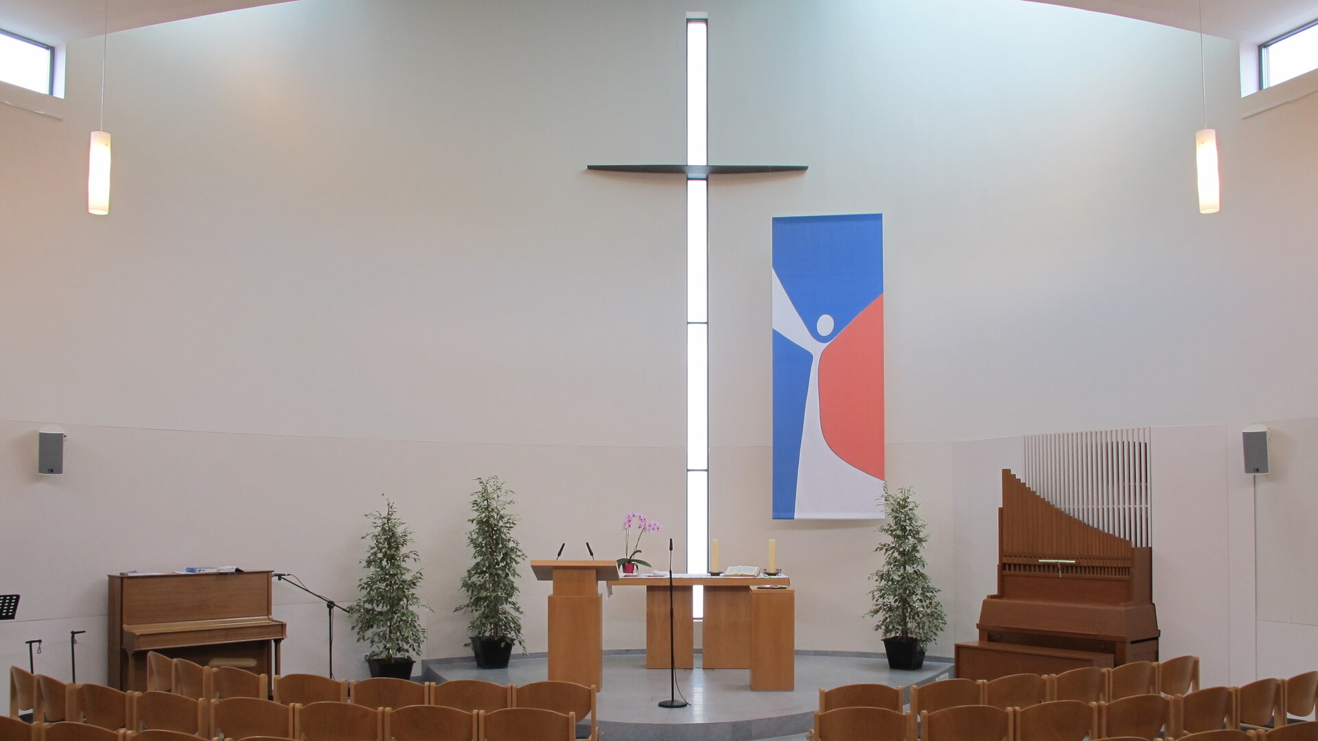 "Evangelischer Gottesdienst - In seiner Spur": Blick in den hellen,  modernen Innenraum der Evangelisch-methodistischen Kirche in Mainz-Kostheim: Die Wände sind weiß, das Gestühl, der Altar und das Klavier sind in hellbraunem Holz gehalten. Ein großes, schlichtes Kreuz befindet sich an der Wand hinter dem Altar, daneben hängt ein abstraktes Gemälde in blau-weiß-rot.