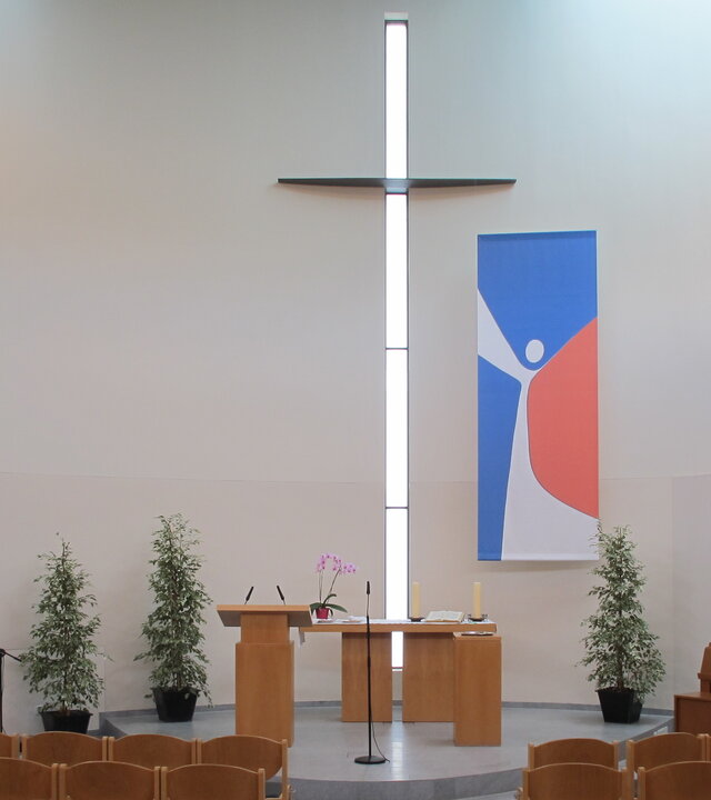 "Evangelischer Gottesdienst - In seiner Spur": Blick in den hellen,  modernen Innenraum der Evangelisch-methodistischen Kirche in Mainz-Kostheim: Die Wände sind weiß, das Gestühl, der Altar und das Klavier sind in hellbraunem Holz gehalten. Ein großes, schlichtes Kreuz befindet sich an der Wand hinter dem Altar, daneben hängt ein abstraktes Gemälde in blau-weiß-rot.