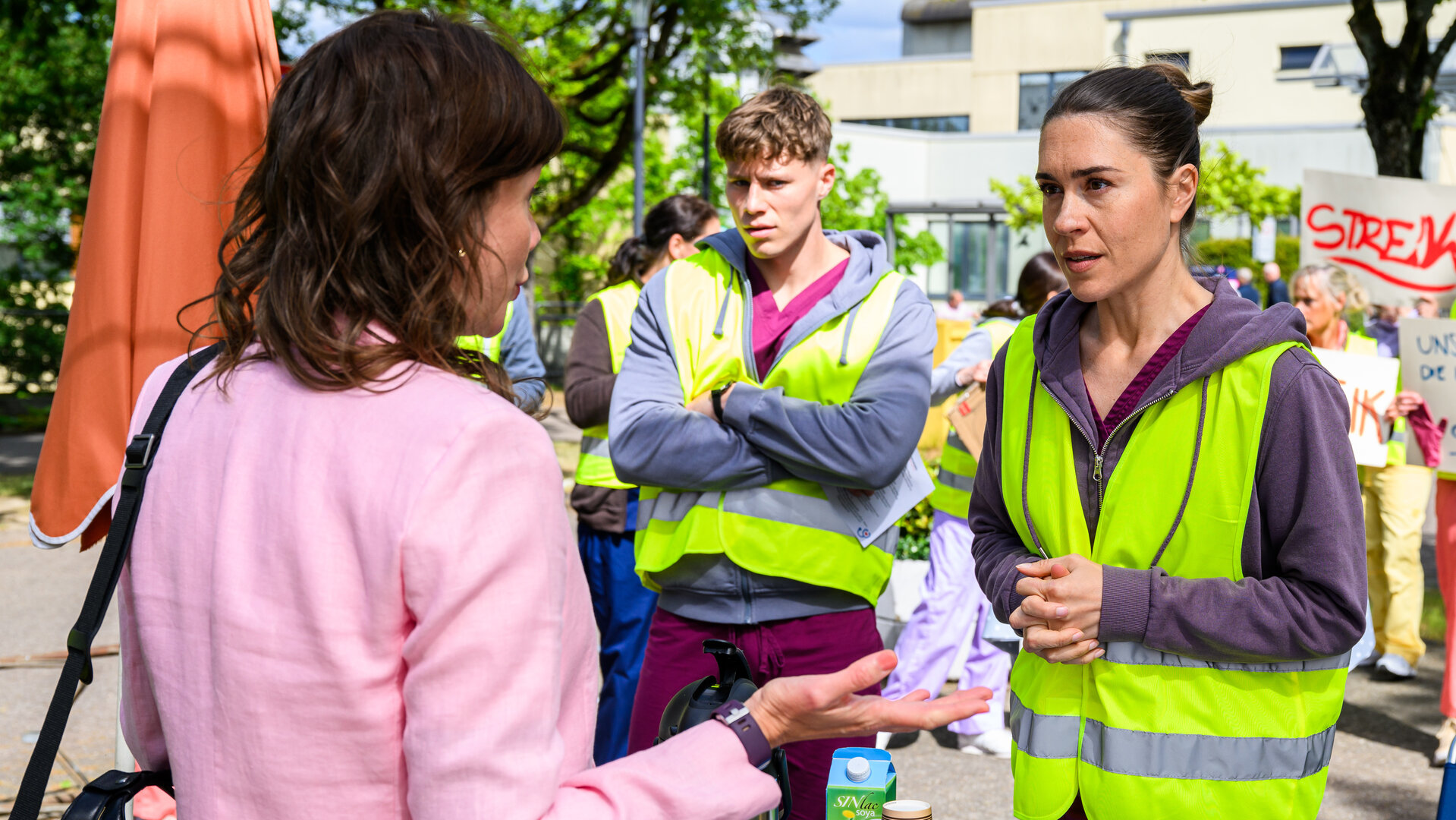 "Bettys Diagnose - Protest": Betty (Henrike Hahn) und Ivo (Oscar Nadermann) stehen im Freien vor Streikenden (Komparsen) an einem Tisch. Ihnen gegenüber steht Katrin Rissmann (Eva Luca Klemmt), sehen sich an.