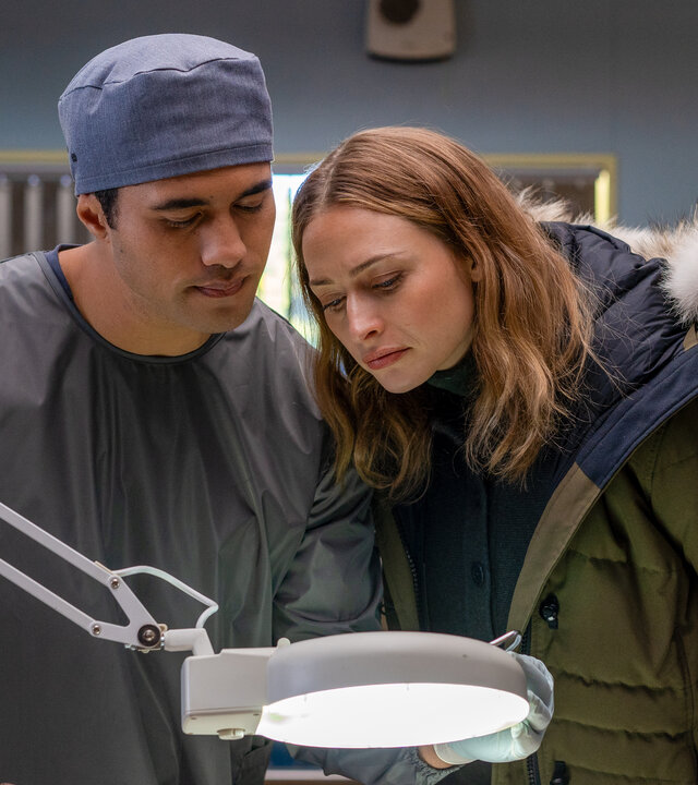 "Queenstown Murders – Zum Sterben schön - Ein explosives Geheimnis": Dr. Ihaka Cooper (Alex Tarrant) und Detective Sergeant Anais Mallory (Chelsie Preston Crayford) stehen eng beieinander an einem Seziertisch. Sie blicken auf einen abgetrennten Arm, der vor ihnen liegt.