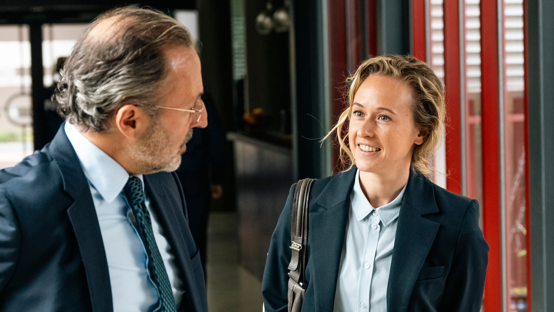 "SOKO Köln - Tod einer Staatsanwältin": Isa Barsch (Nurit Hirschfeld) steht neben Steffen Kehlau (Robert Schupp) im Kommissariat. Der Staatsanwalt hat seinen Kopf zu Isa gedreht und steht seitlich vor der Kamera. Isa sieht ihn fröhlich an.