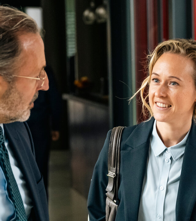 "SOKO Köln - Tod einer Staatsanwältin": Isa Barsch (Nurit Hirschfeld) steht neben Steffen Kehlau (Robert Schupp) im Kommissariat. Der Staatsanwalt hat seinen Kopf zu Isa gedreht und steht seitlich vor der Kamera. Isa sieht ihn fröhlich an.