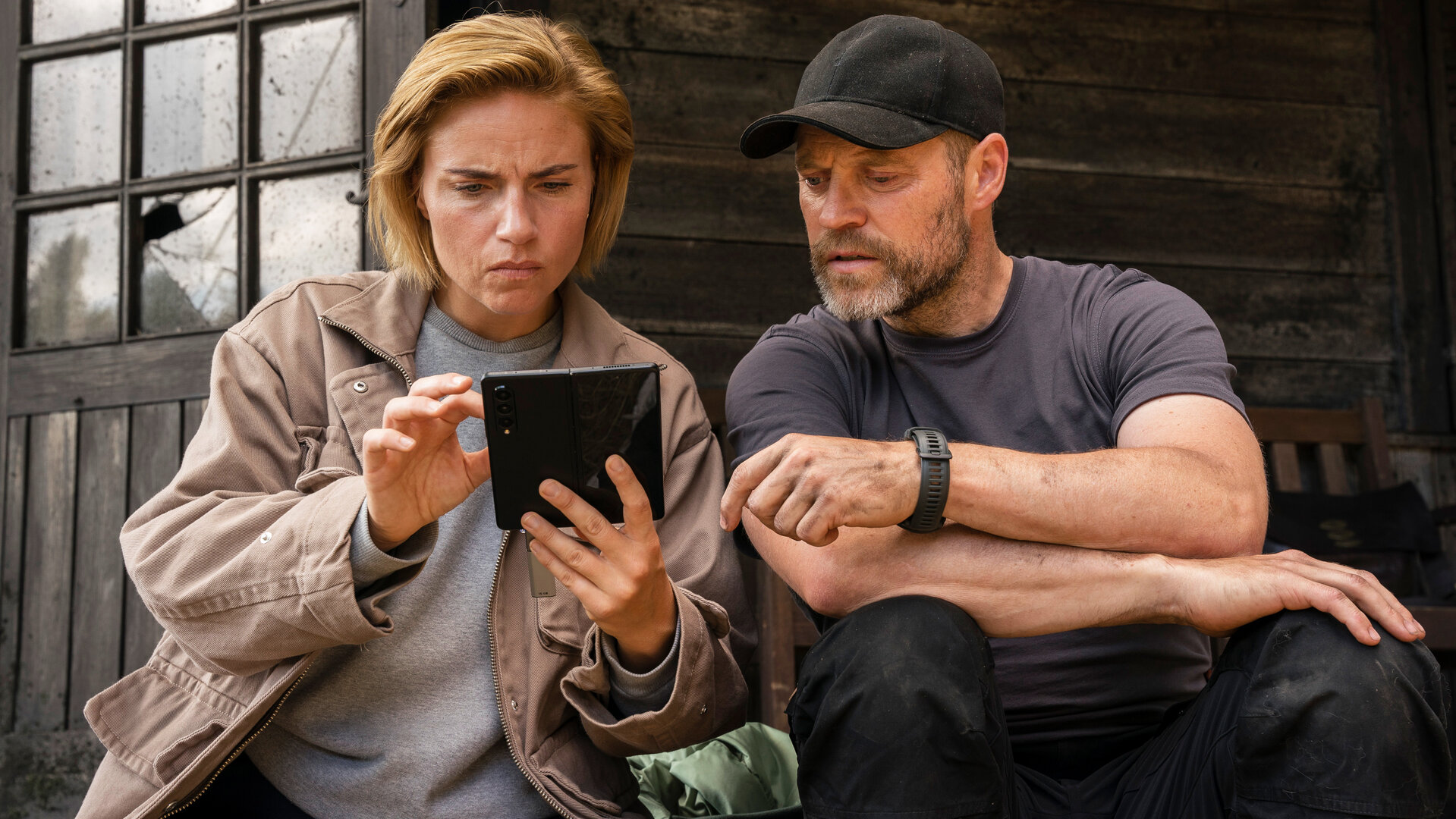 "Oxen - Versteckte Gegner": Margrethe (Josephine Park) und Niels (Jacob Lohmann) sitzen nebeneinander vor einer Holzhütte. Sie schauen auf ein aufgeklapptes Smartphone.