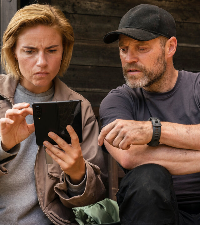 "Oxen - Versteckte Gegner": Margrethe (Josephine Park) und Niels (Jacob Lohmann) sitzen nebeneinander vor einer Holzhütte. Sie schauen auf ein aufgeklapptes Smartphone.